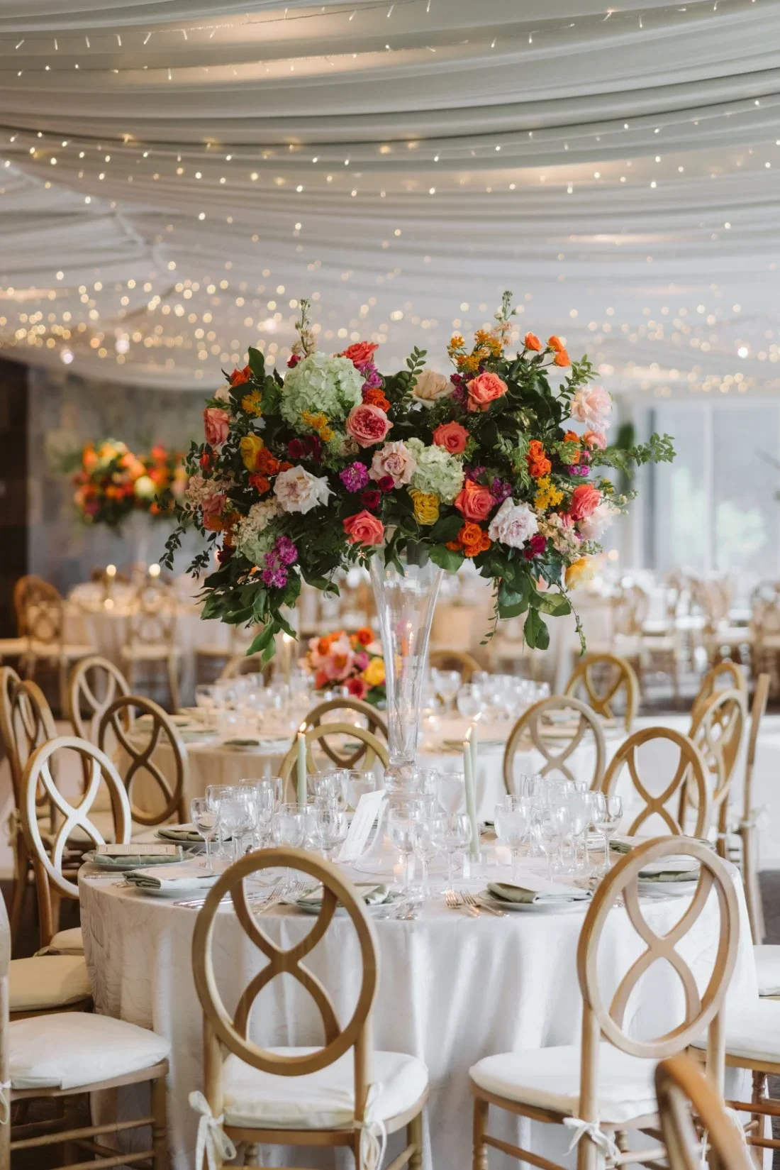 Wedding lighting ideas featuring delicate twinkle lights across a draped reception ceiling above colorful floral centerpieces and elegant gold chairs.