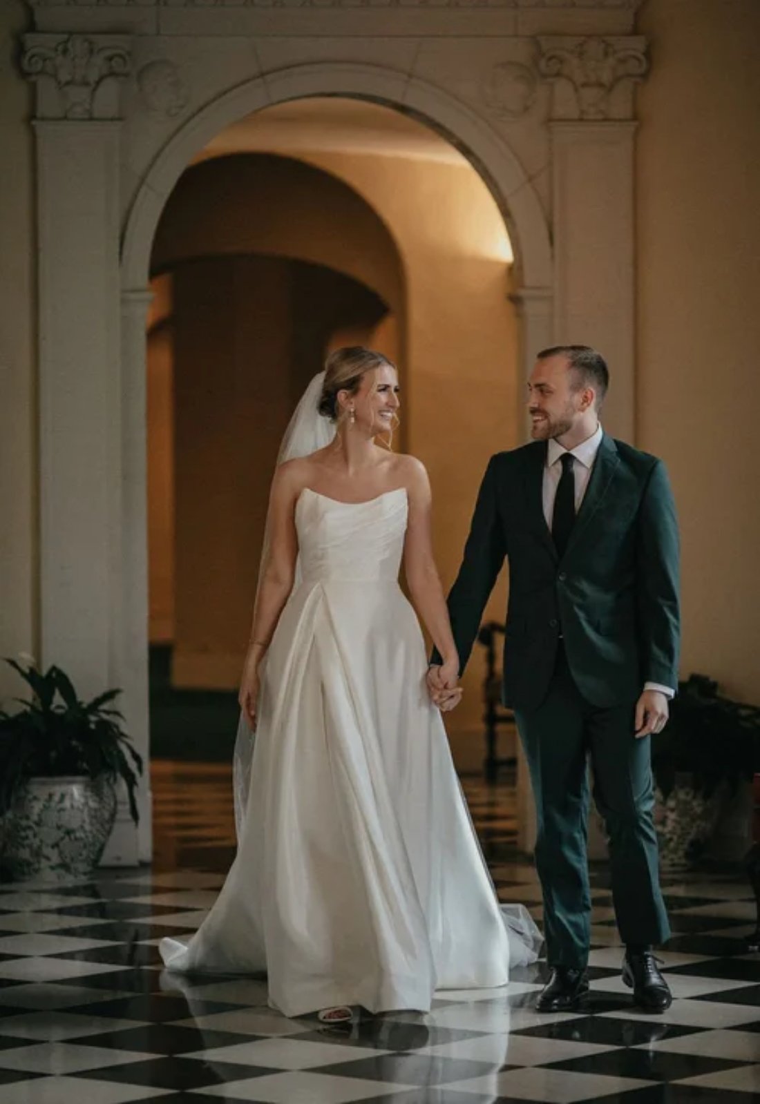 A bride in a strapless gown walking hand in hand with her groom in a dark green suit inside a historic venue, highlighting timeless winter wedding fashion for couples.