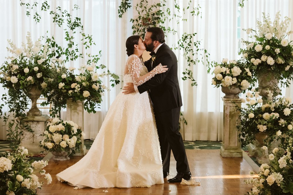 Bride and groom sharing a kiss after their indoor wedding ceremony surrounded by timeless white floral arrangements.