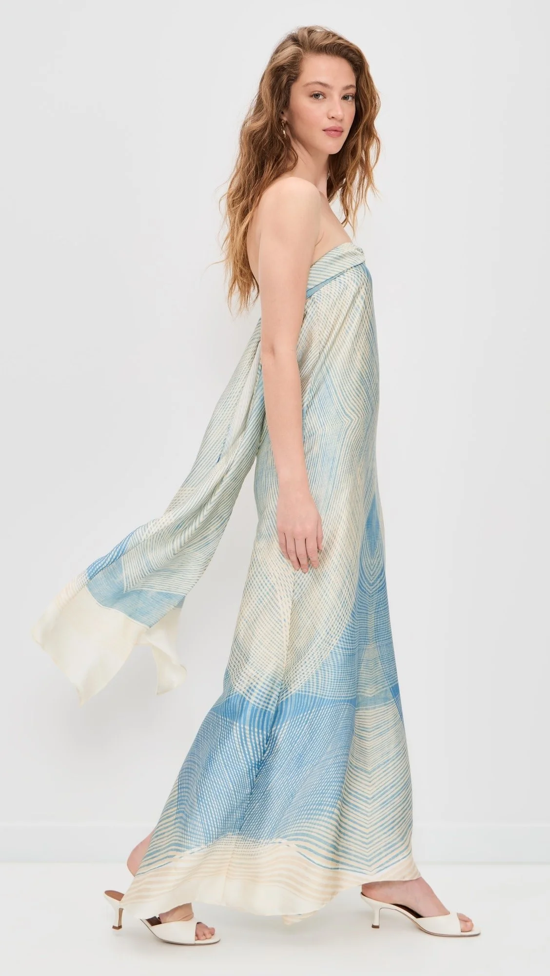 A woman with long wavy brown hair wearing a strapless, flowing maxi dress with blue, beige, and white patterns, and white heeled sandals, standing against a plain light gray background.