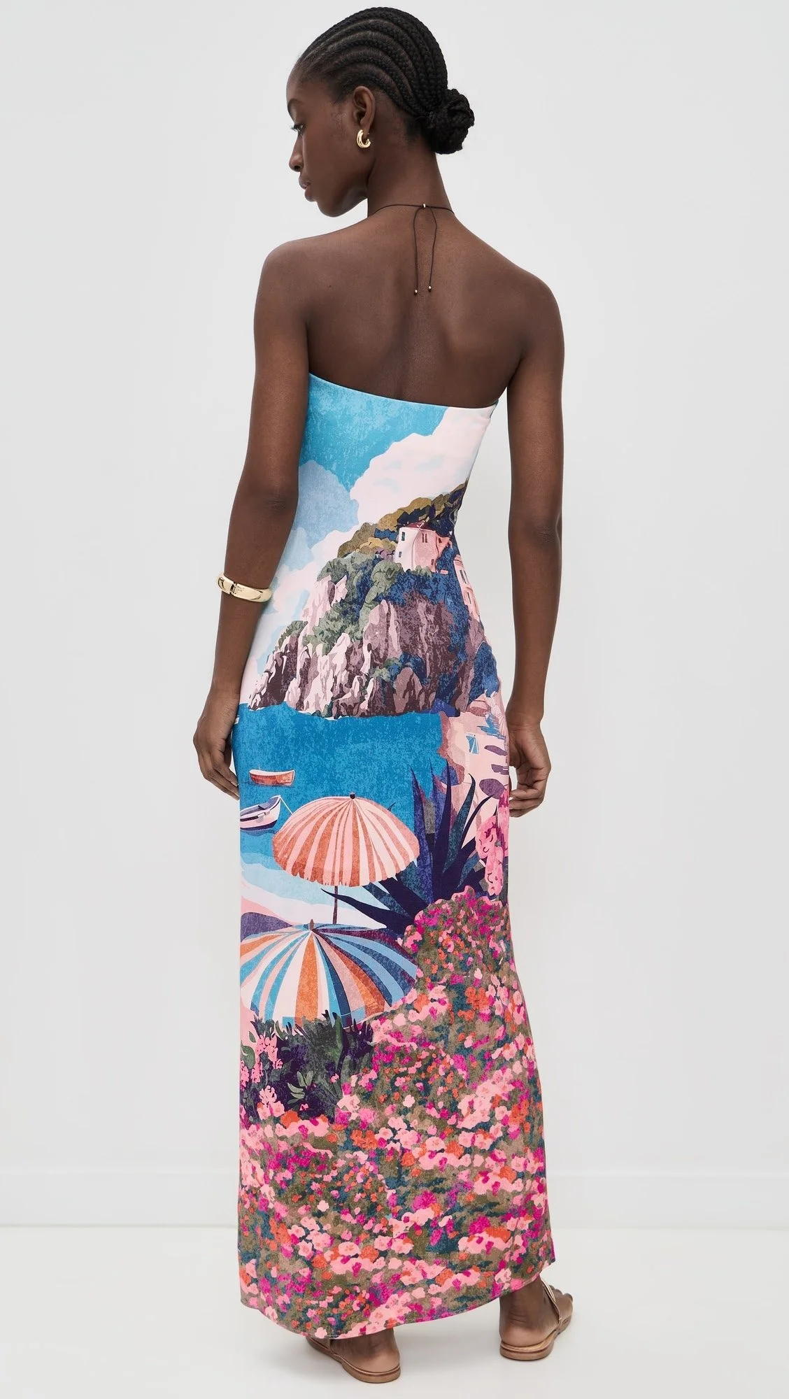 A woman wearing a strapless dress with a scenic beach and mountain landscape print, standing sideways against a plain white background.