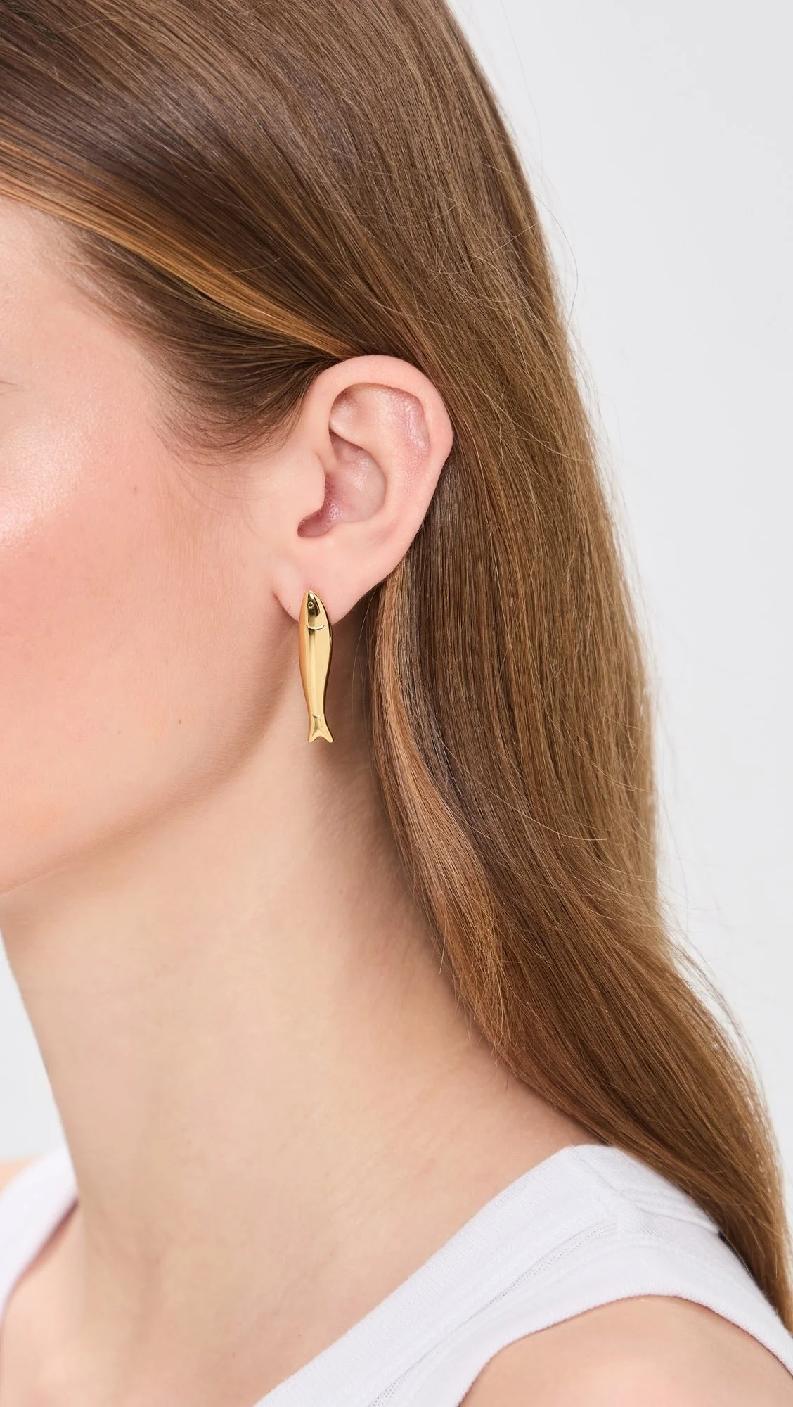 Close-up of a woman's ear with a gold fish-shaped earring, with red hair and a white sleeveless top.