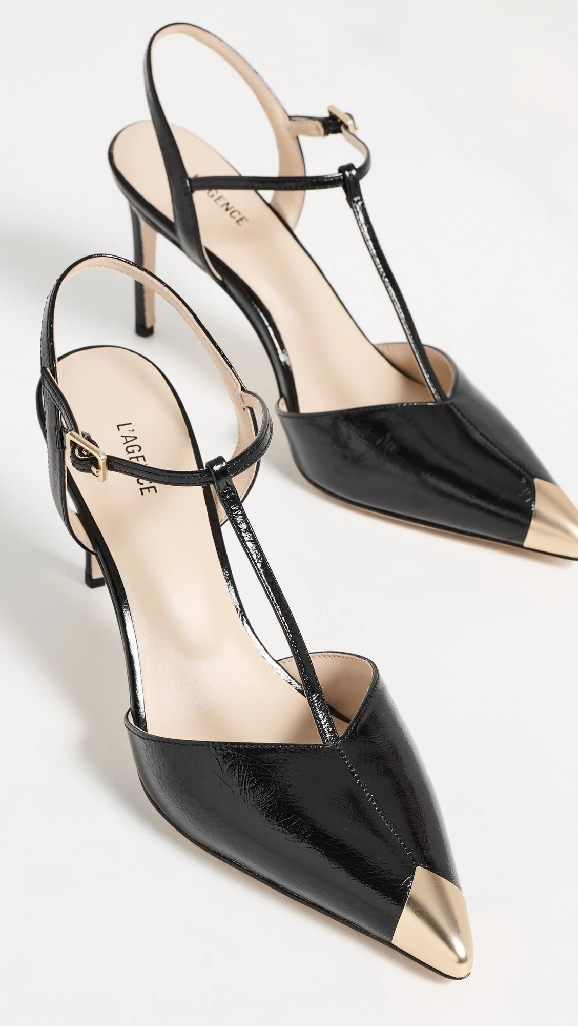 A pair of black high-heeled shoes with pointed beige and silver toes, ankle straps, and stiletto heels on a white background.