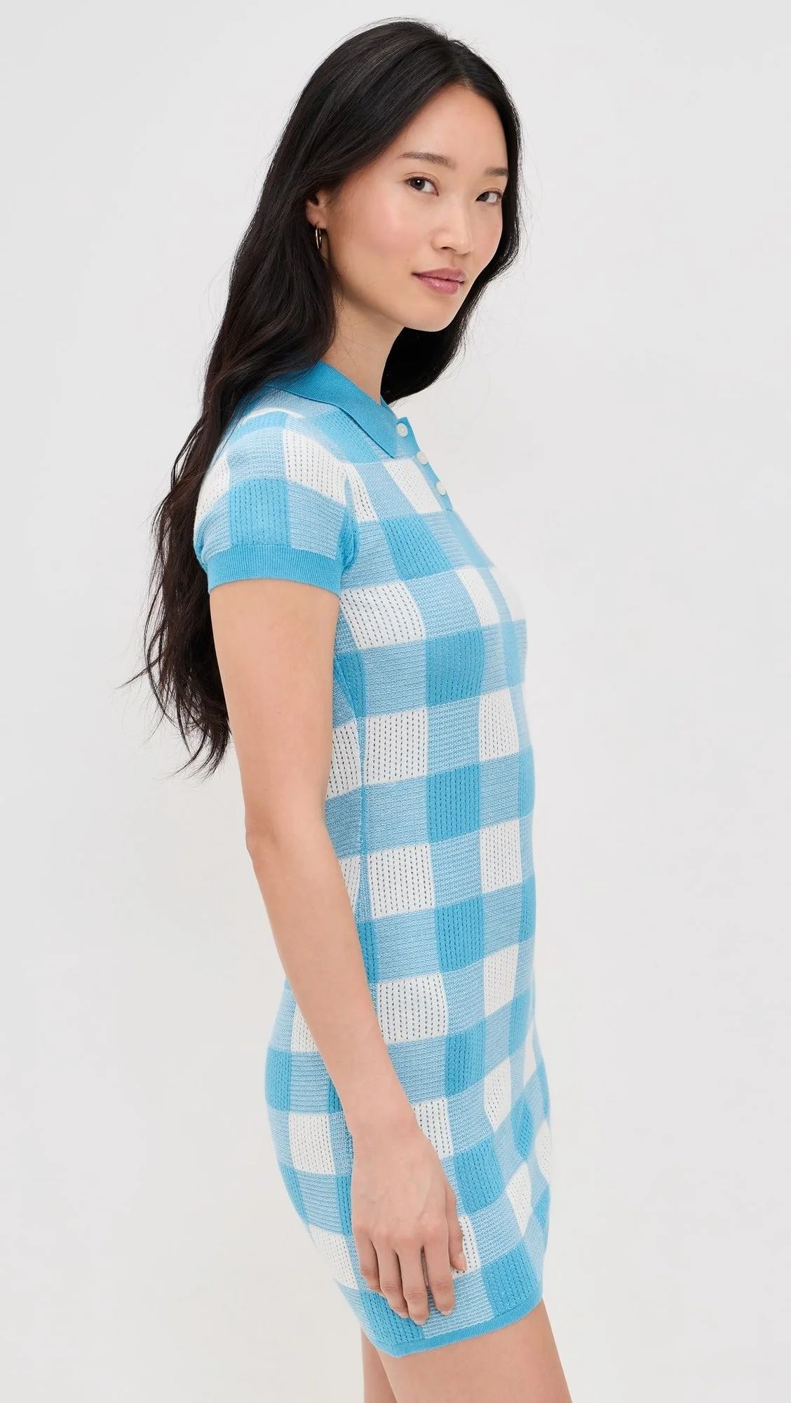 A woman with long dark hair wearing a blue and white checkered short-sleeve knit dress with a collar and button placket, standing against a plain white background.