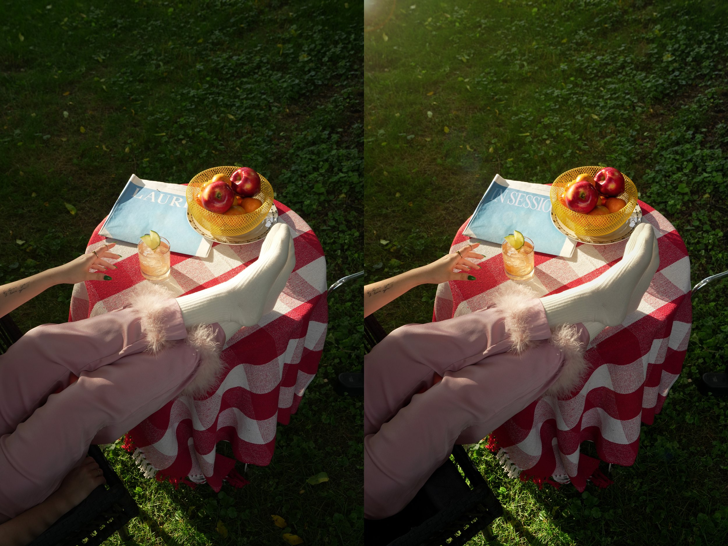 A person with green nail polish holding a glass of iced beverage with a lemon slice, on a red and white checkered tablecloth, with a yellow basket of apples and oranges, a blue magazine, and a white cloth on a table outdoors on grass.