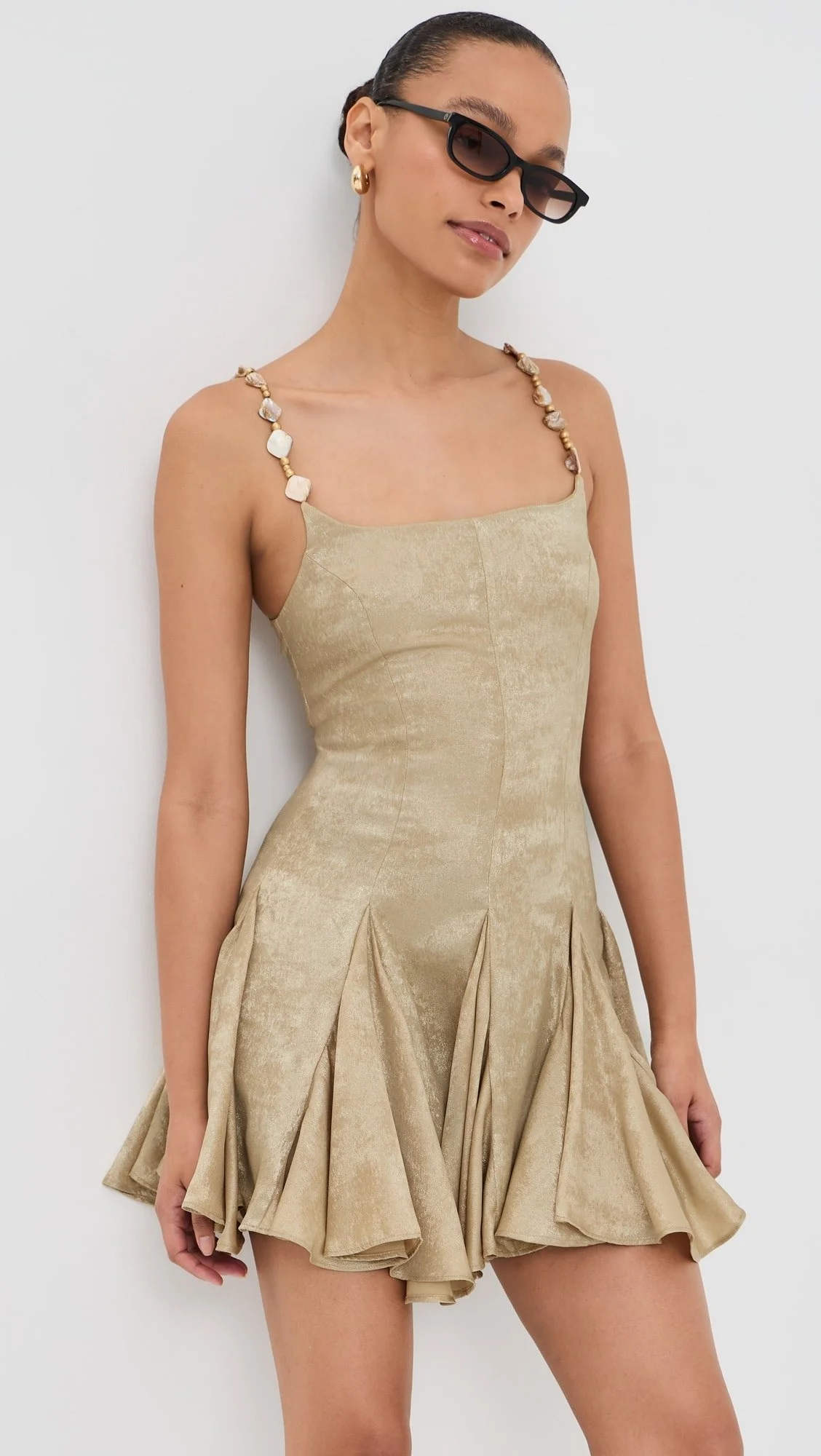 A woman wearing a beige, satin-like dress with a pleated skirt, black sunglasses, gold earrings, and a shell necklace, standing against a plain white background.