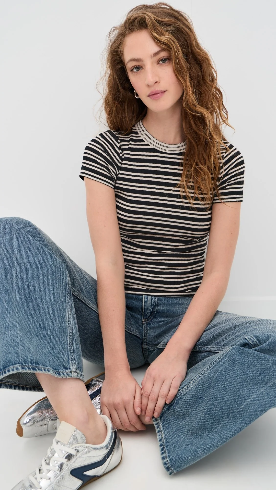 A young woman with wavy reddish-brown hair wearing a black and white striped t-shirt, blue jeans, and silver sneakers, sitting with her legs crossed and hands clasped in front of her against a plain white background.