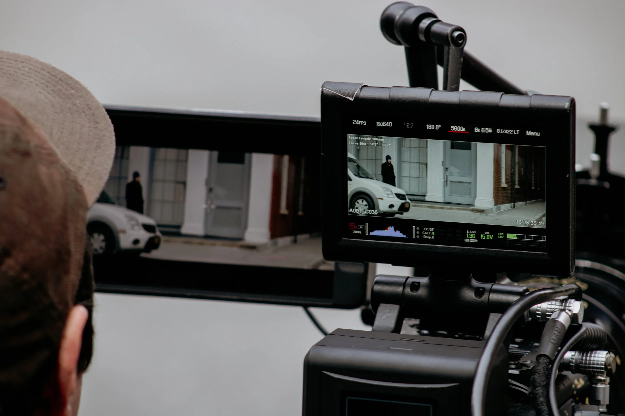 A camera monitor showing a scene with a white car and person standing outside a building, with a person wearing a hat and ear protection in the foreground.