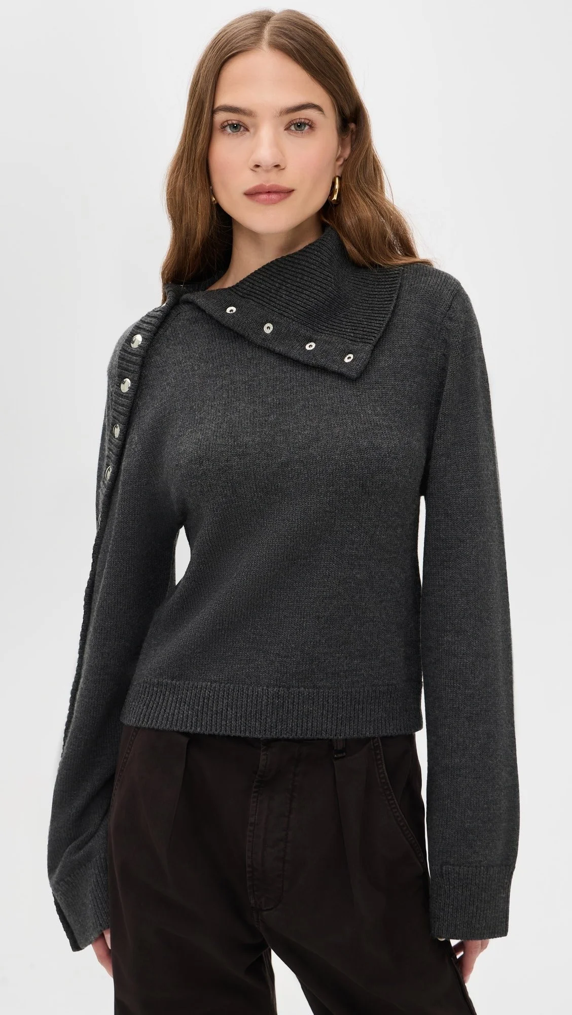 A woman with long brown hair wearing a dark gray sweater with metallic button details on the shoulder and a collar, paired with black pants.