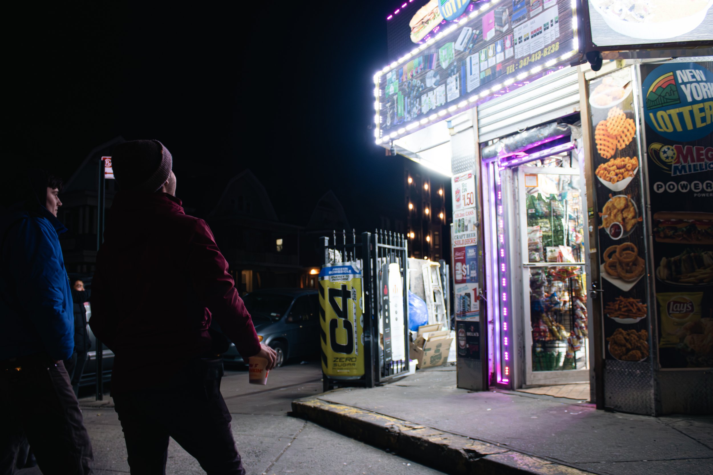 Two people standing in front of a brightly lit food stand at night, with one holding a drink, outside a fair or carnival setting.