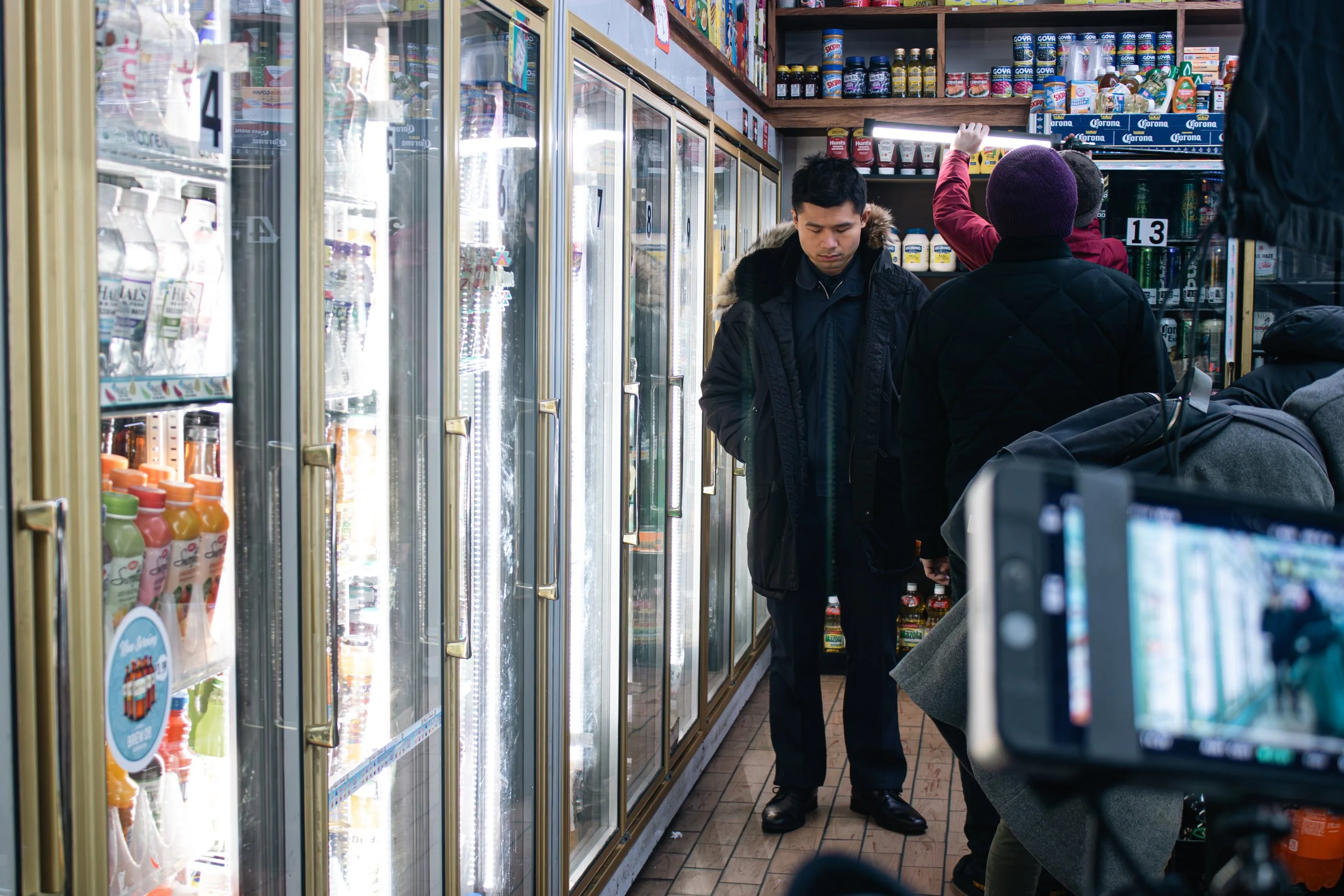 Two people shopping at a grocery store aisle with refrigerated drinks. One person is wearing a black jacket with a fur-lined hood and is looking down, while the other, in a purple knit hat and black jacket, is reaching towards the top shelf.