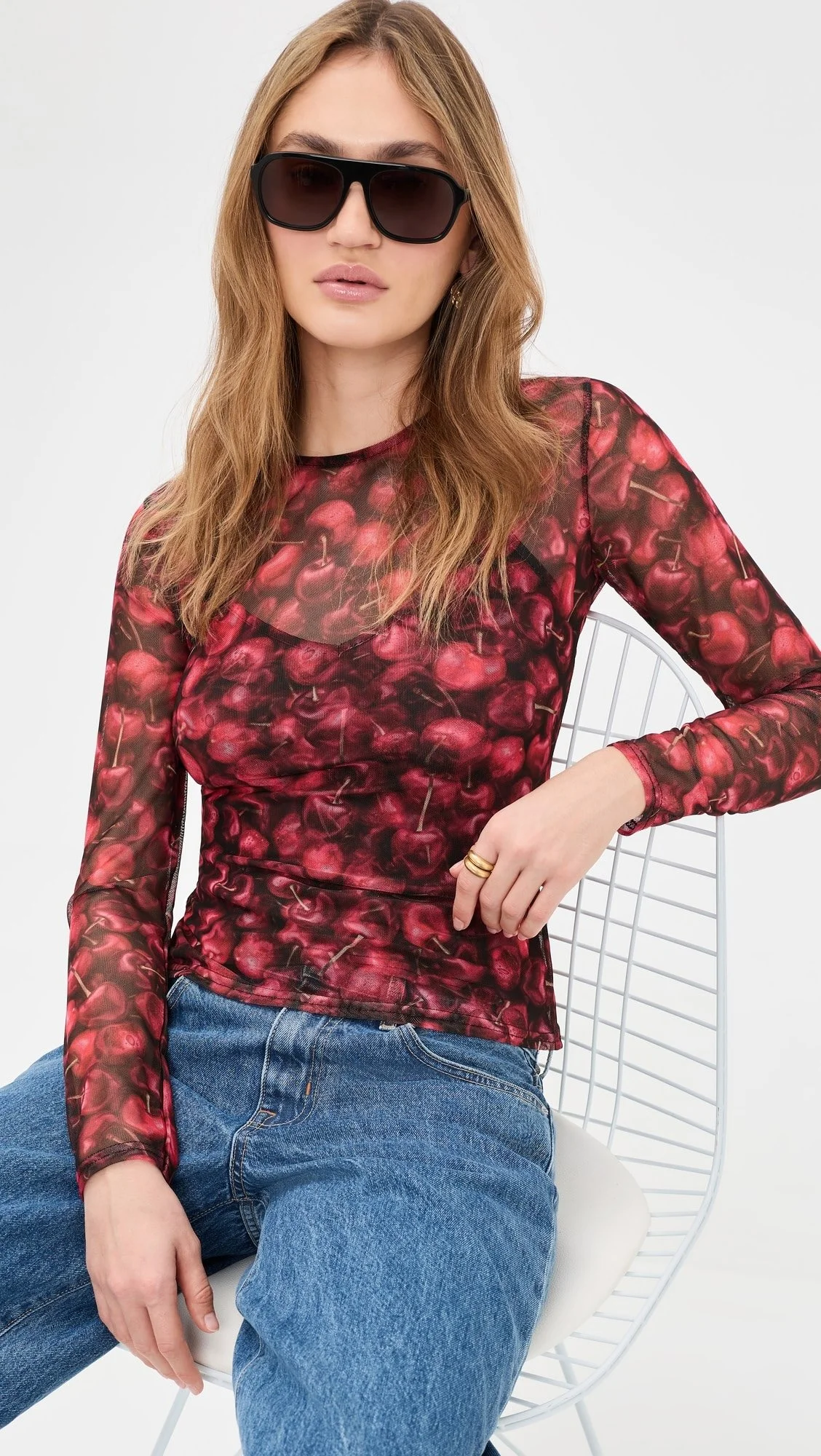 A woman with long wavy auburn hair wearing large black sunglasses, a cherry-patterned long sleeve top, and blue jeans, sitting on a white wire chair.