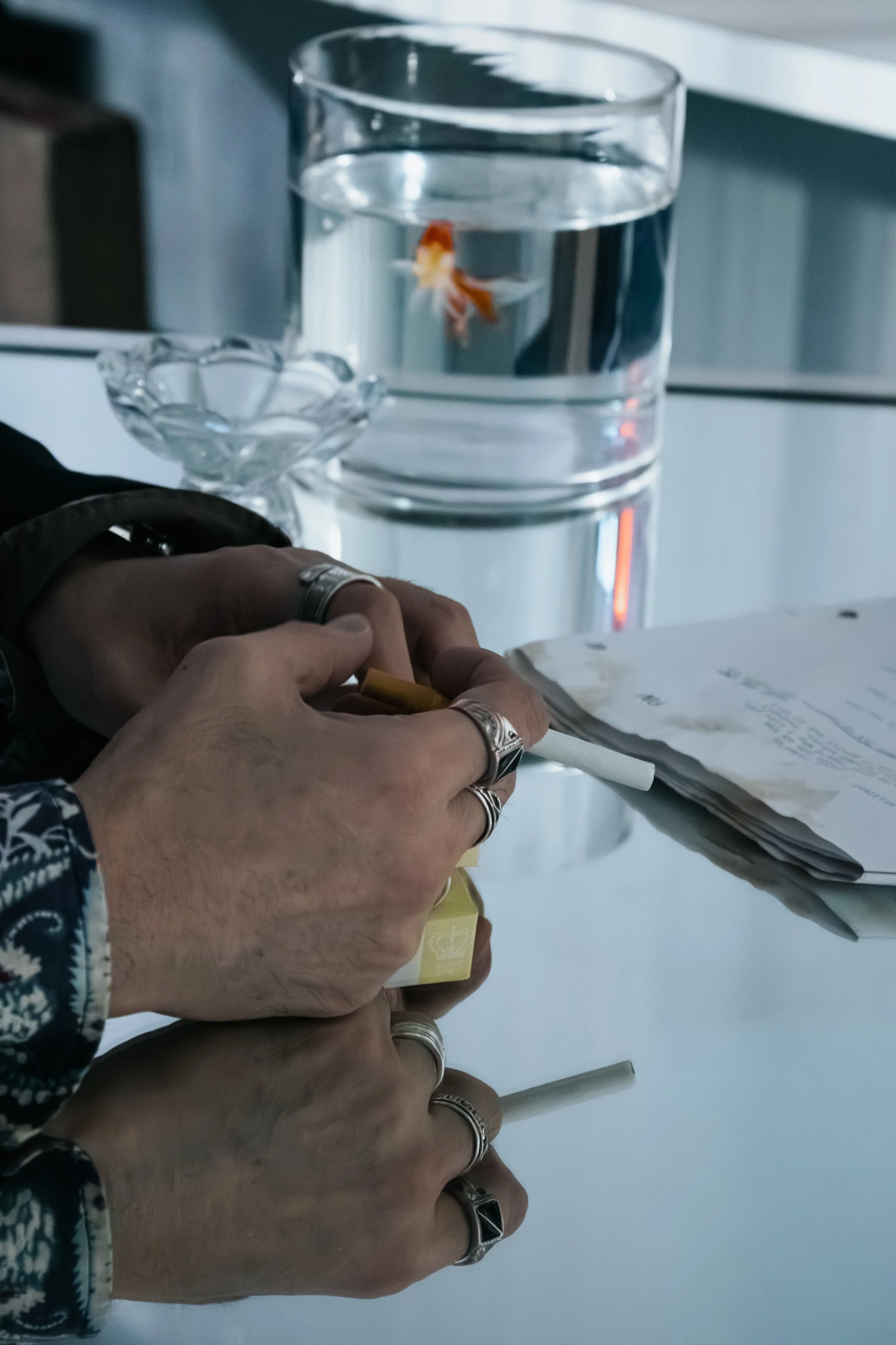 Two hands on a table with a glass of water and a fishbowl in the background containing a goldfish, and some papers and objects on the table.