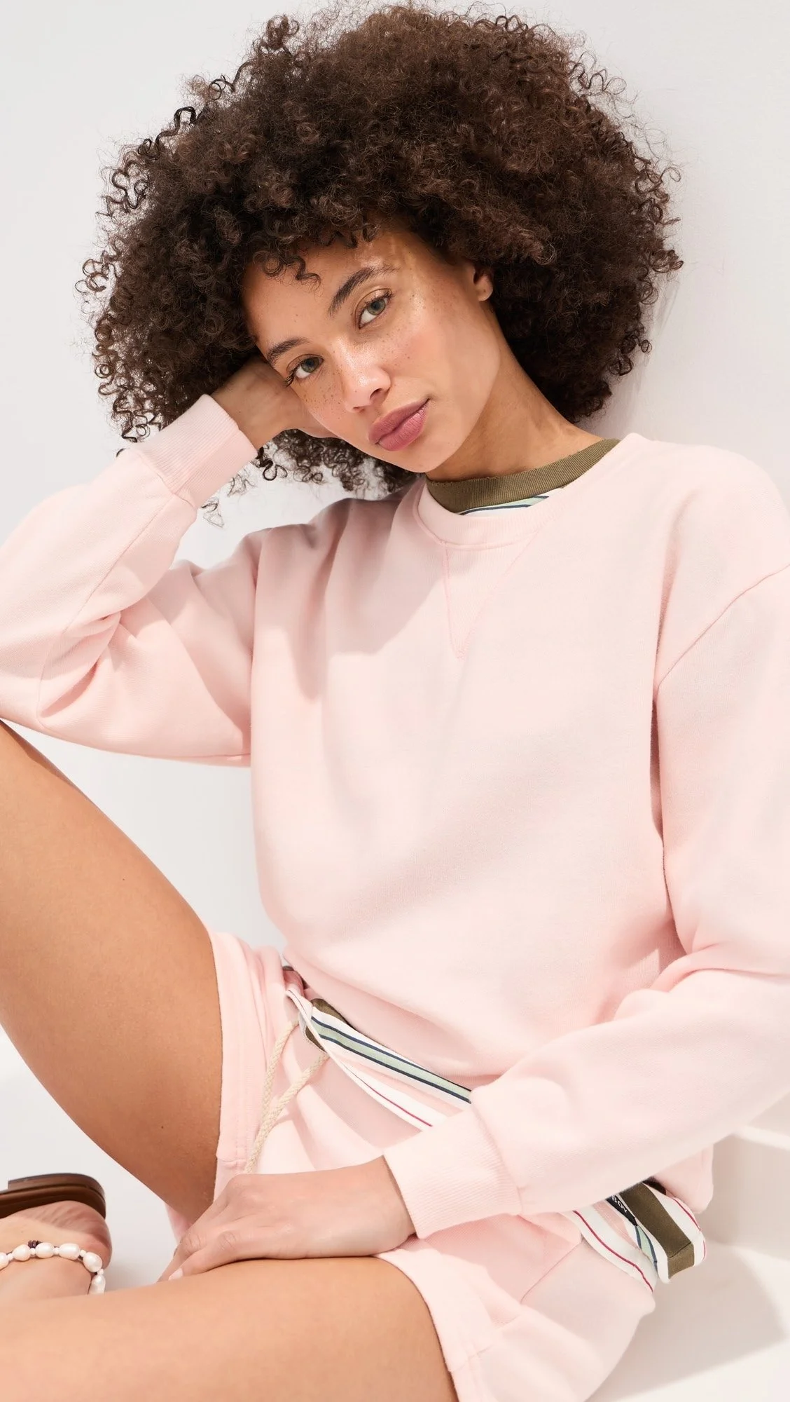 Young woman with curly hair, wearing a pink sweatshirt, posing with her arm resting on her head, against a plain white background.