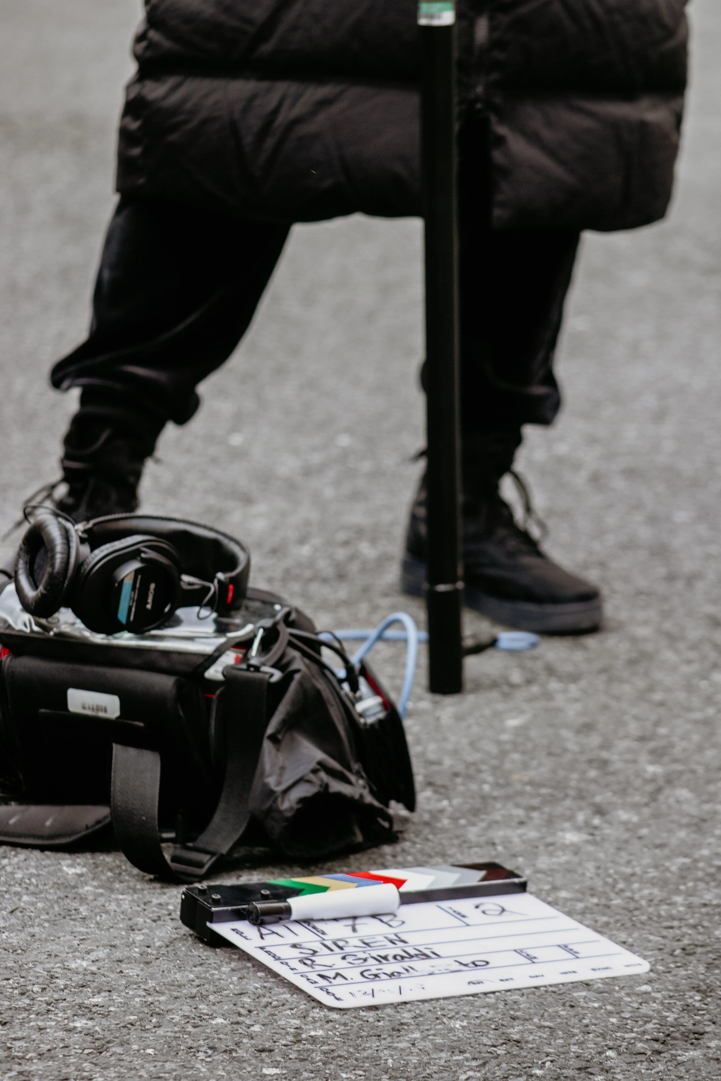 Close-up of a person's lower body, wearing black pants and shoes, standing behind recording equipment on a gray asphalt ground. There is a black bag with headphones on top and a whiteboard with handwritten text in the foreground.