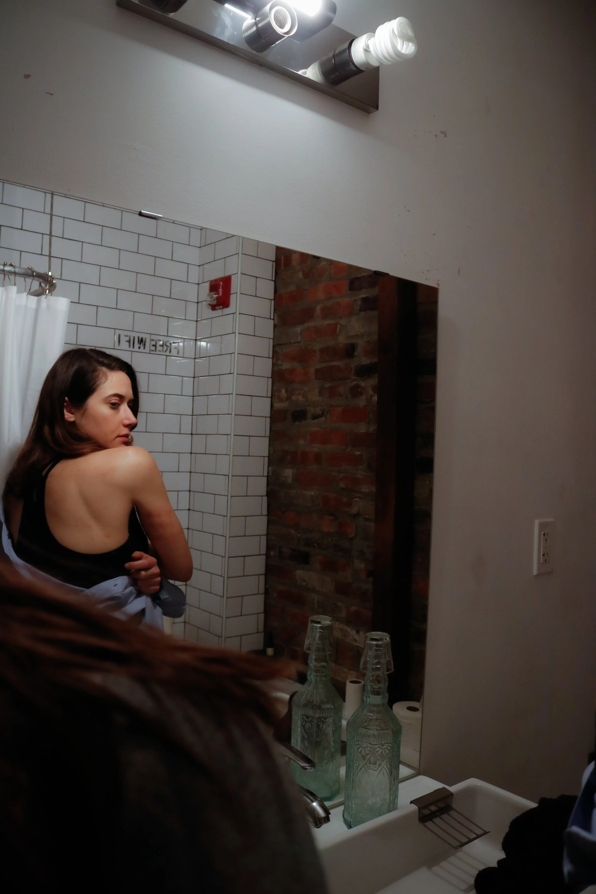A woman with brown hair looking over her shoulder in a bathroom with white subway tiles, a brick wall, and a mirror. The bathroom countertop has glass bottles and a sink with a drying rack, and the background shows a shower area with a white curtain 