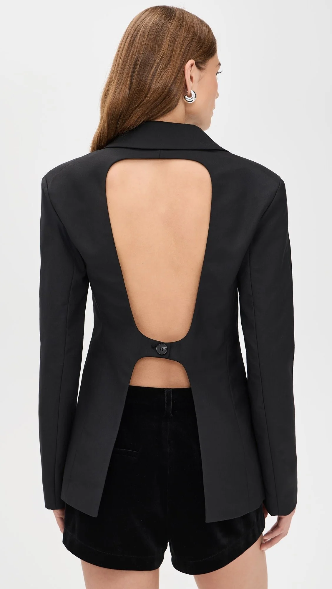 A woman with brown hair wearing a black blazer with a large cutout back and black shorts, standing against a plain white background.