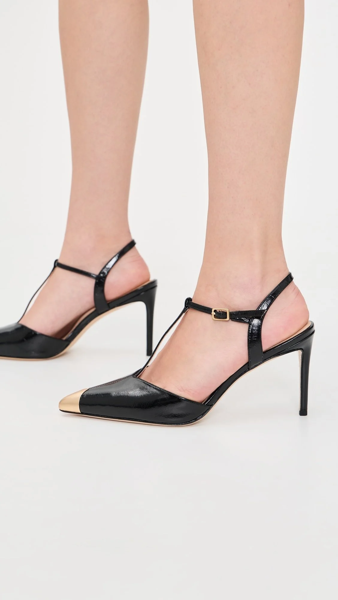Close-up of a woman wearing black high-heeled shoes with pointed toes and ankle straps, standing on a white background.