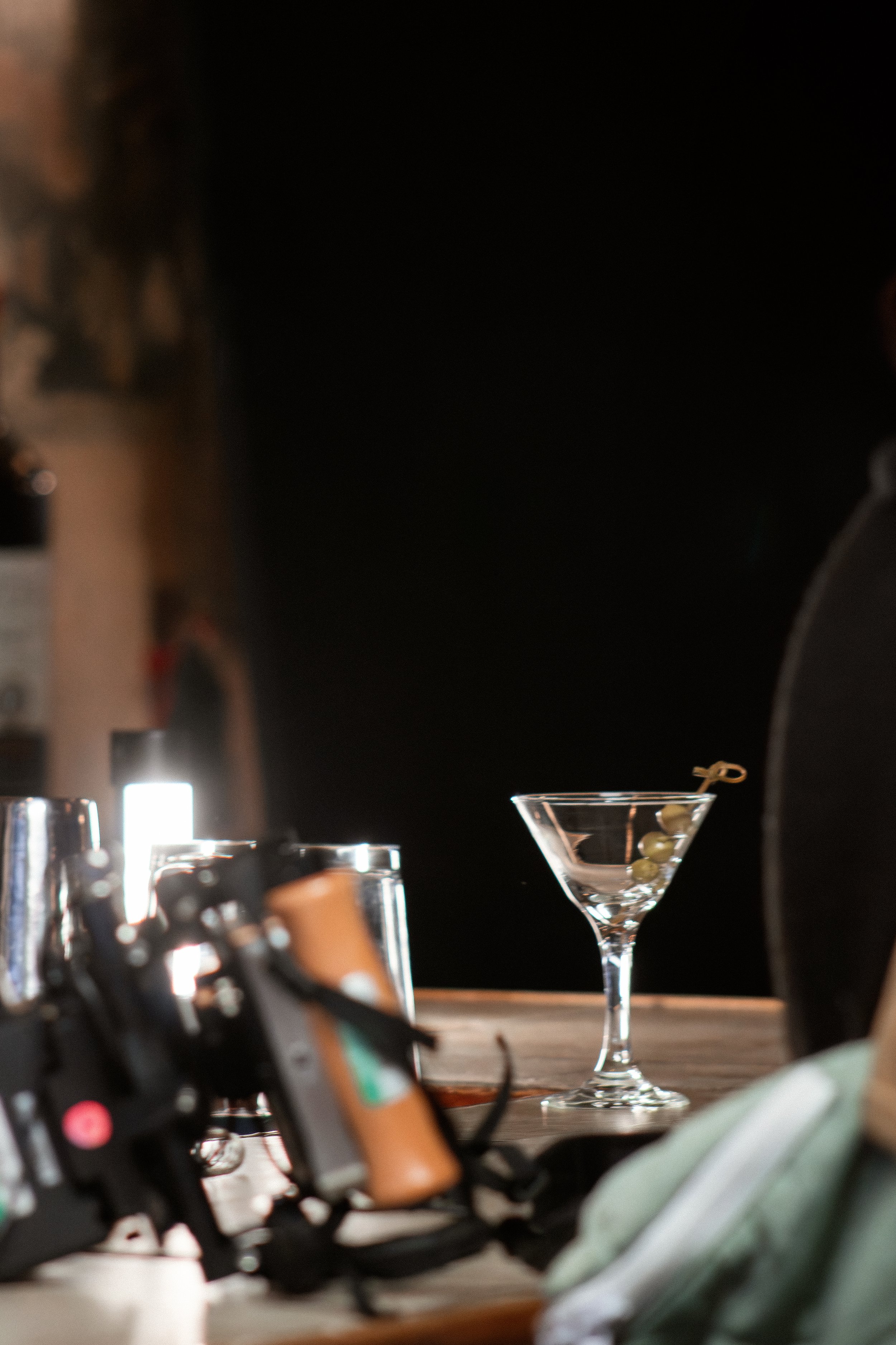 A bar counter with various bottles and bar tools in the foreground. In the background, there is an empty martini glass with four green olives on a skewer inside. The setting suggests a bar or cocktail lounge.