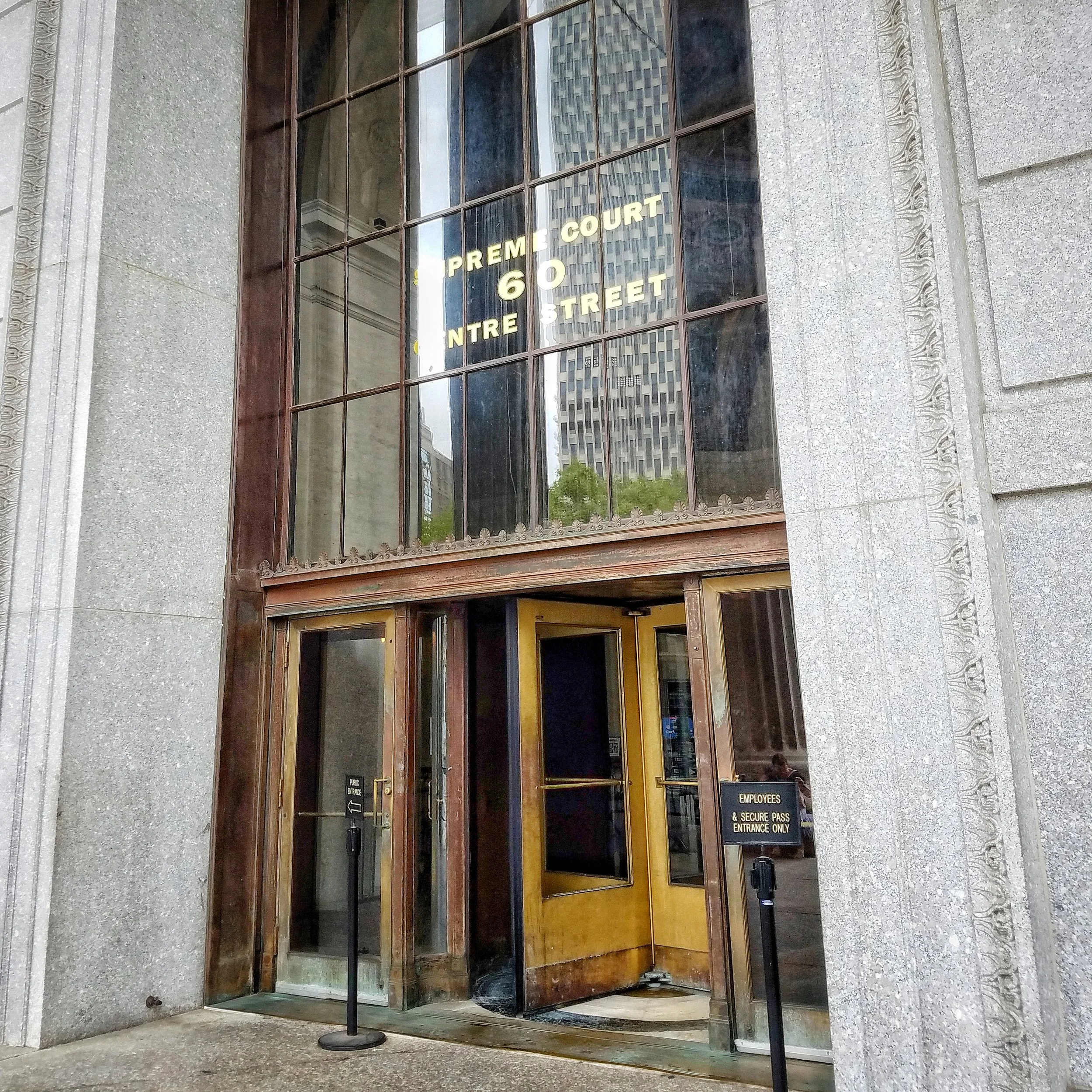 The entrance to 60 Centre shows the building’s address in the windows (“Supreme Court 60 Centre Street”). There is a wooden revolving door in the center and two regular doors on either side. The regular door on the right is reserved for employees.