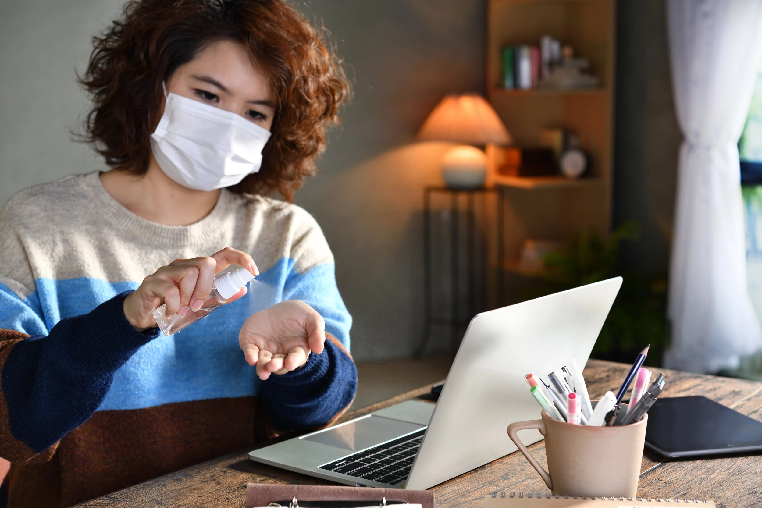  Woman in quarantine and social distancing wearing surgical mask and cleaning hands with alcohol gel sanitizer while working from home during Covid-19 Coronavirus pandemic 