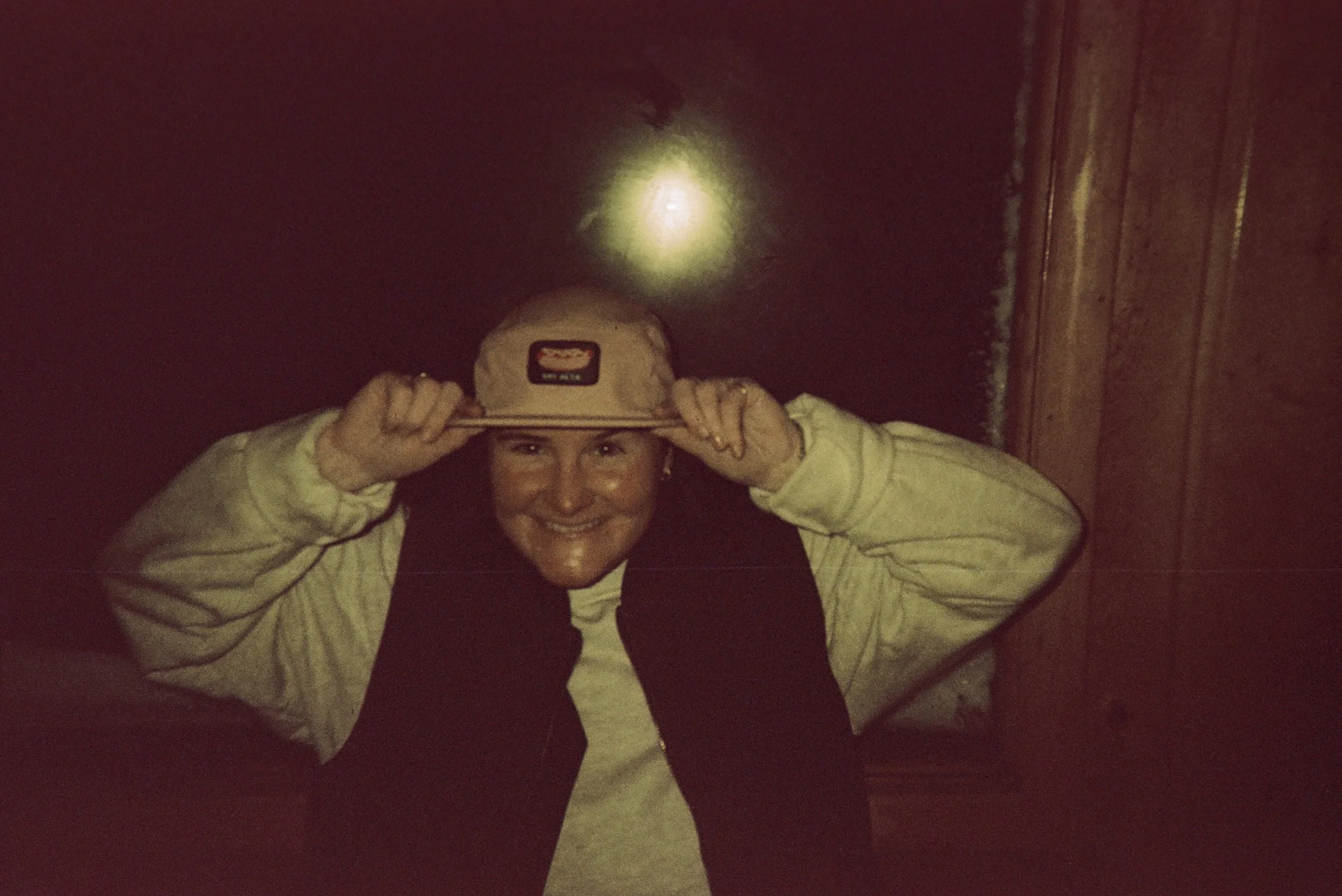 A person with short hair and smiling face, wearing a beige and black jacket, holding a baseball cap with both hands above their head, standing against a dark wall with a bright light above.