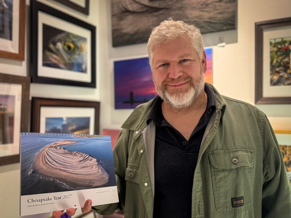 Photographer Bill Mish holding the Chesapeake Year 2027 calendar in front of a wall of his framed photographs