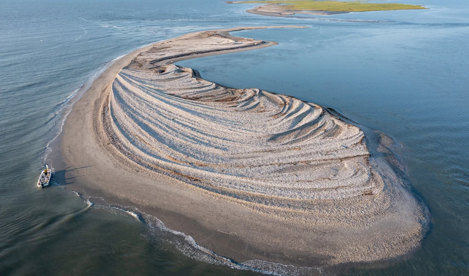 Aerial photograph of a curving barrier-island sand spit ringed by shallow water, with a small boat pulled up at its edge — the cover of Chesapeake Year 2027