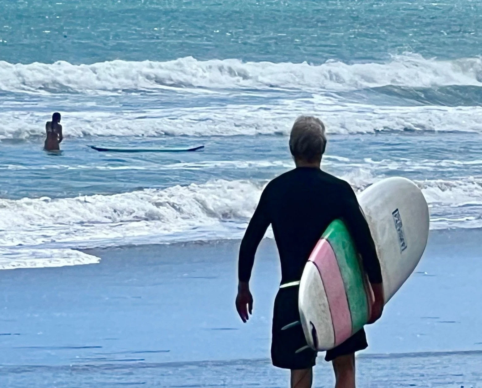Man walking on the beach holding a surfboard with a person in the water nearby.