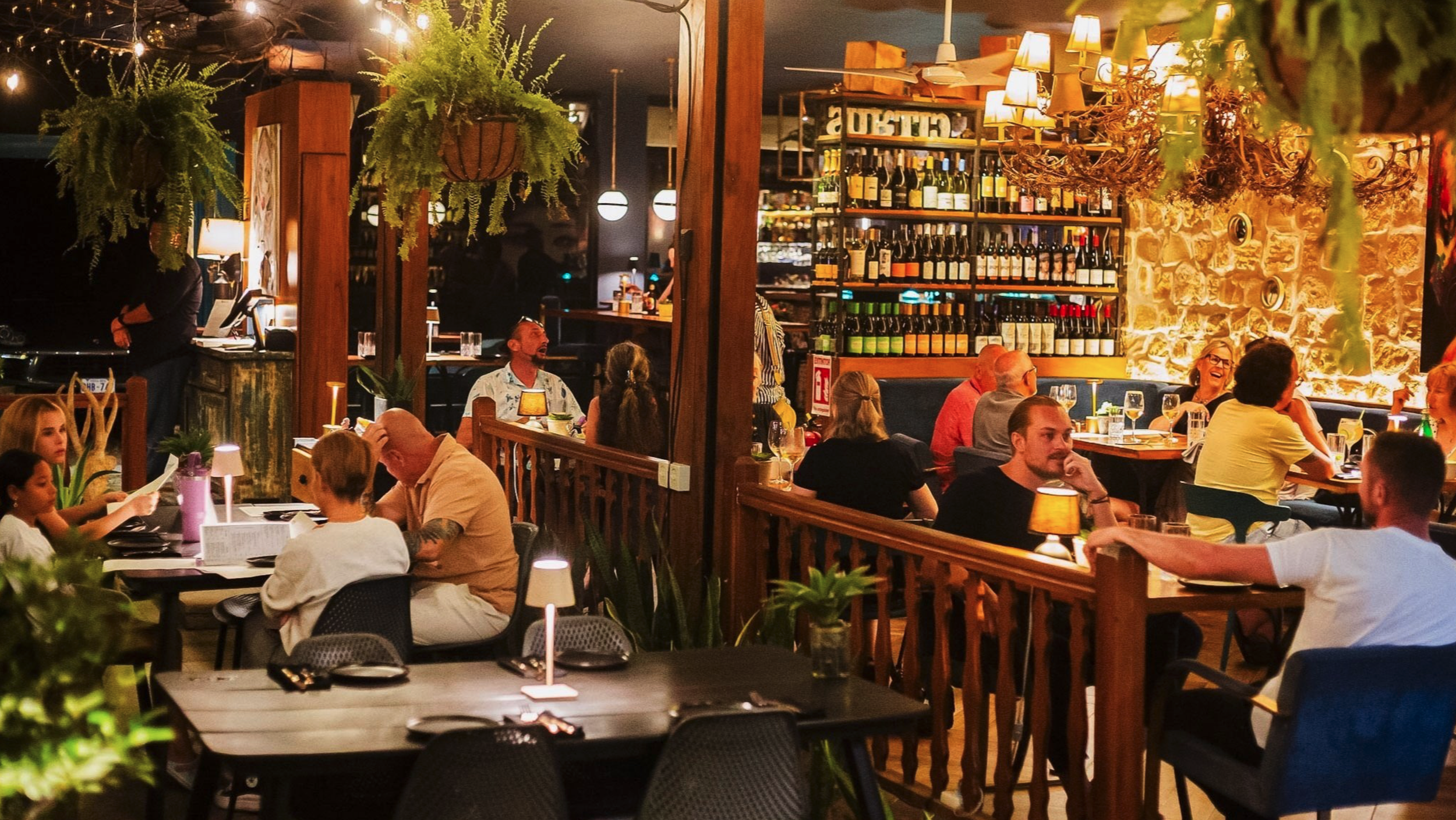 La Junta Dominical is along the river with great food. Cozy restaurant interior with diners, wooden furniture, hanging plants, ambient lighting, and wine bottles on display.