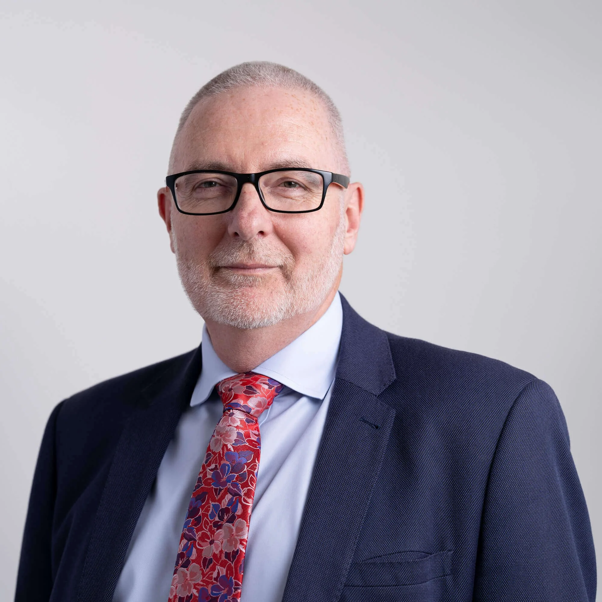 Professional man with glasses and a floral tie wearing a suit, smiling against a plain gray background.