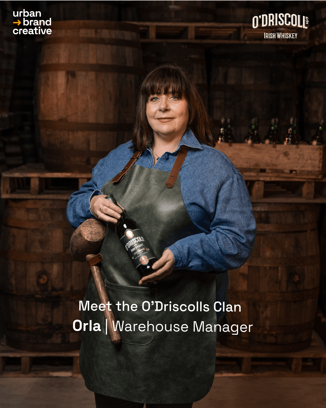 A woman standing in a whiskey warehouse holding a bottle of O'Driscoll's Irish Whiskey. She is wearing a denim shirt and a leather apron, with wooden whiskey barrels in the background.