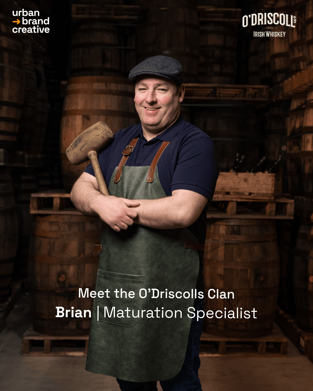 A man smiling in a whiskey cellar surrounded by wooden barrels, holding a hammer, wearing a flat cap, navy polo shirt, and a green apron.
