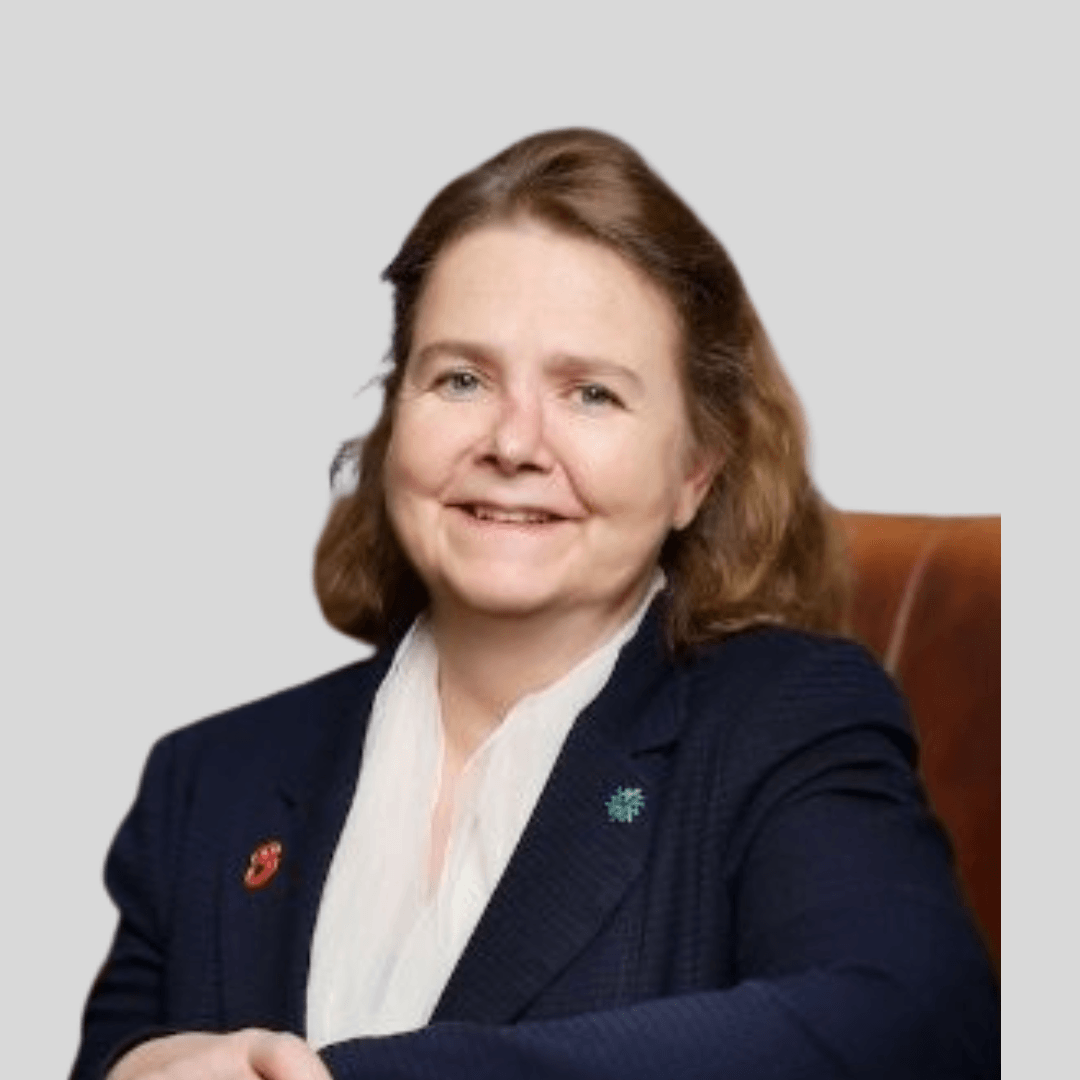 Portrait of a woman with light skin, shoulder-length brown hair, wearing a dark blazer, white blouse, and pins, sitting on a leather chair against a plain gray background.