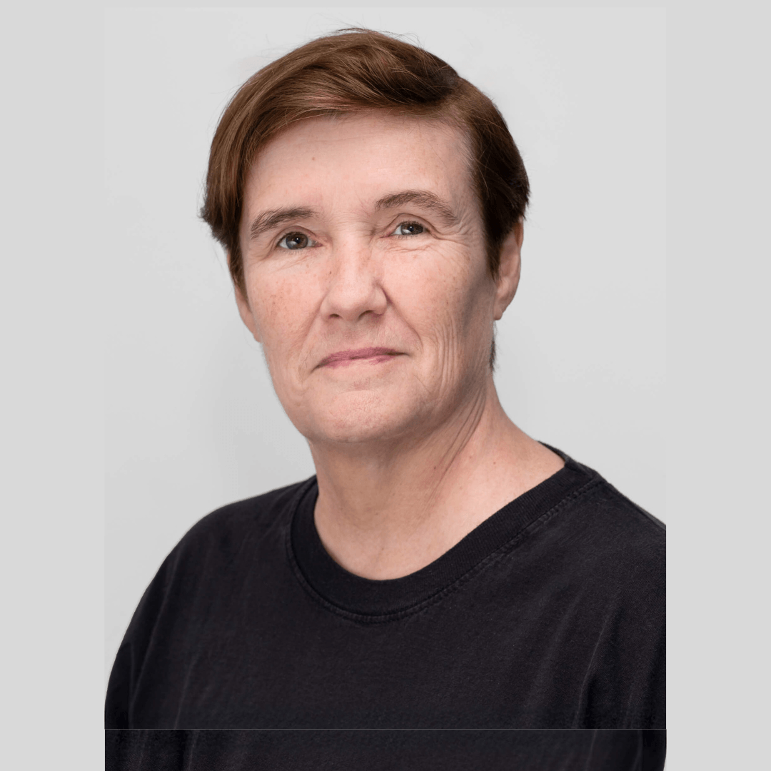 Close-up of a middle-aged woman with short brown hair, wearing a black shirt, against a light gray background.