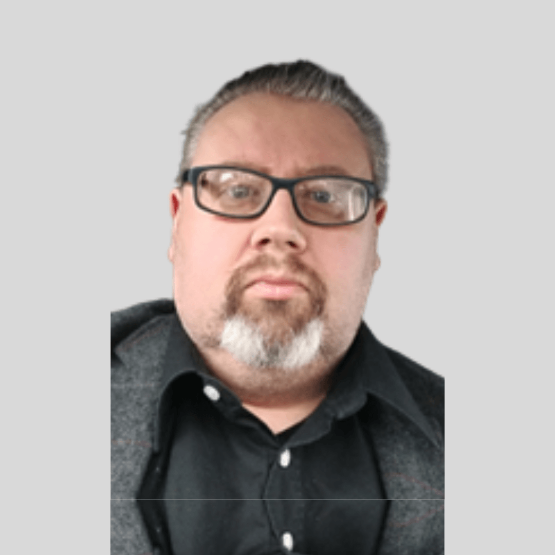 Close-up photo of a man with glasses, short gray hair, and a beard, wearing a black shirt and a gray jacket against a light gray background.