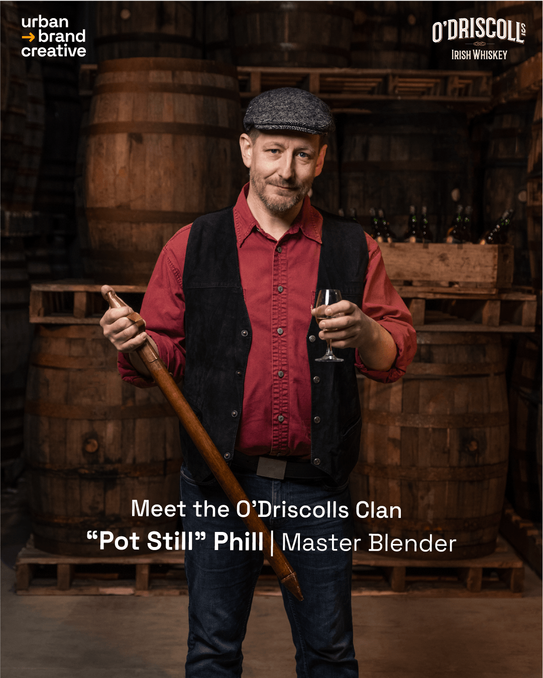 A man in a flat cap, red shirt, and black vest holding a smoking pipe and a glass of whiskey inside a distillery with wooden barrels in the background.