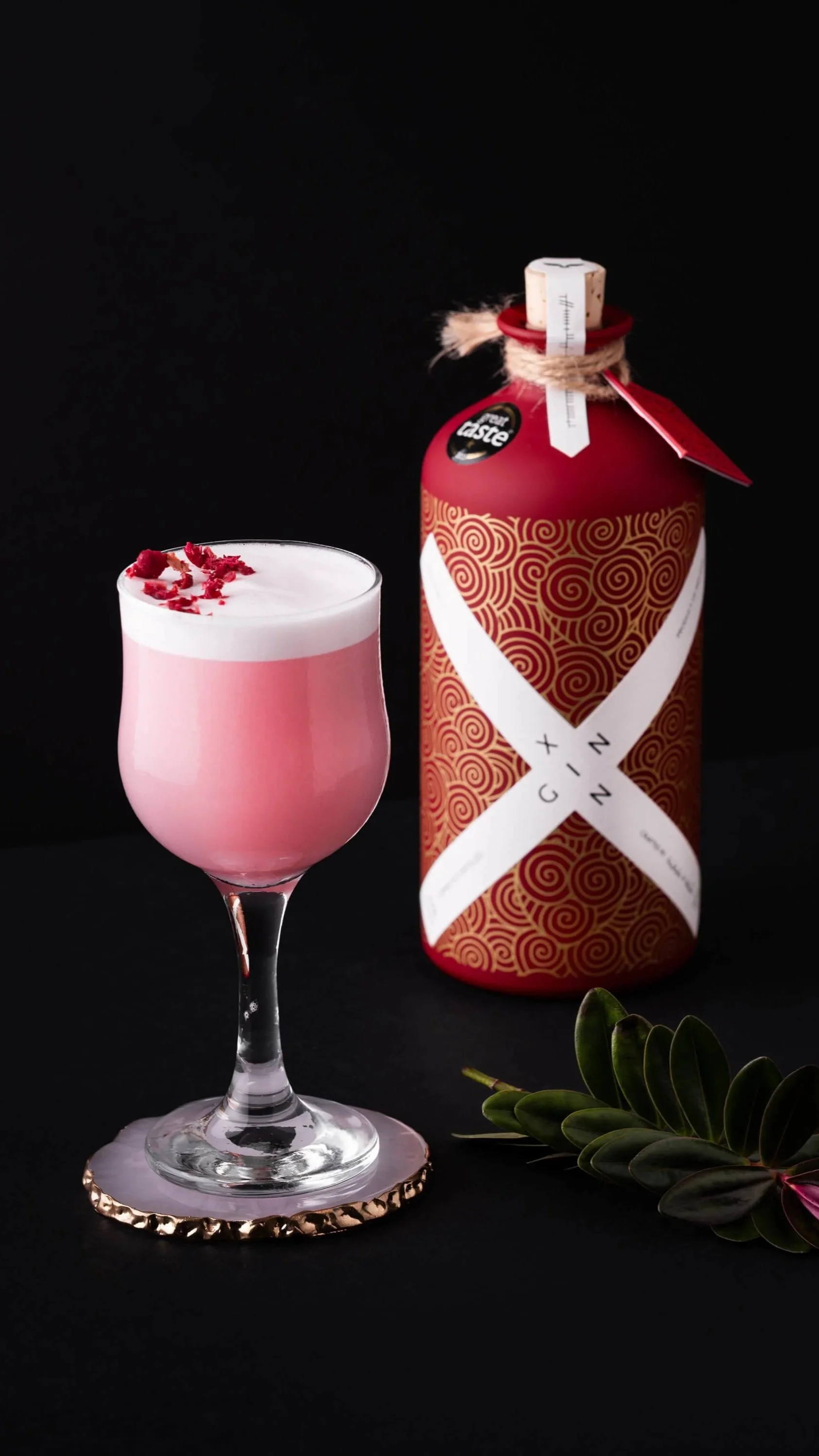 A pink cocktail in a stemmed glass garnished with red dried flower petals, set on a decorative coaster, with a red and gold spiral-patterned bottle labeled 'Gin' and a green leafy plant beside it, all against a black background.