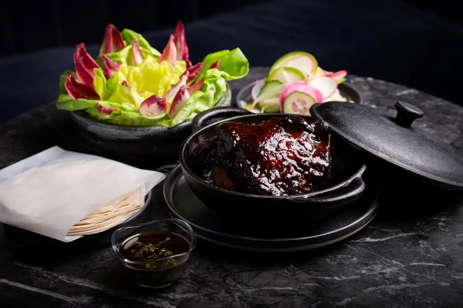 A plate of cooked meat with sauce, fresh mixed greens, sliced radishes and a small bowl of dipping sauce on a dark marble surface.