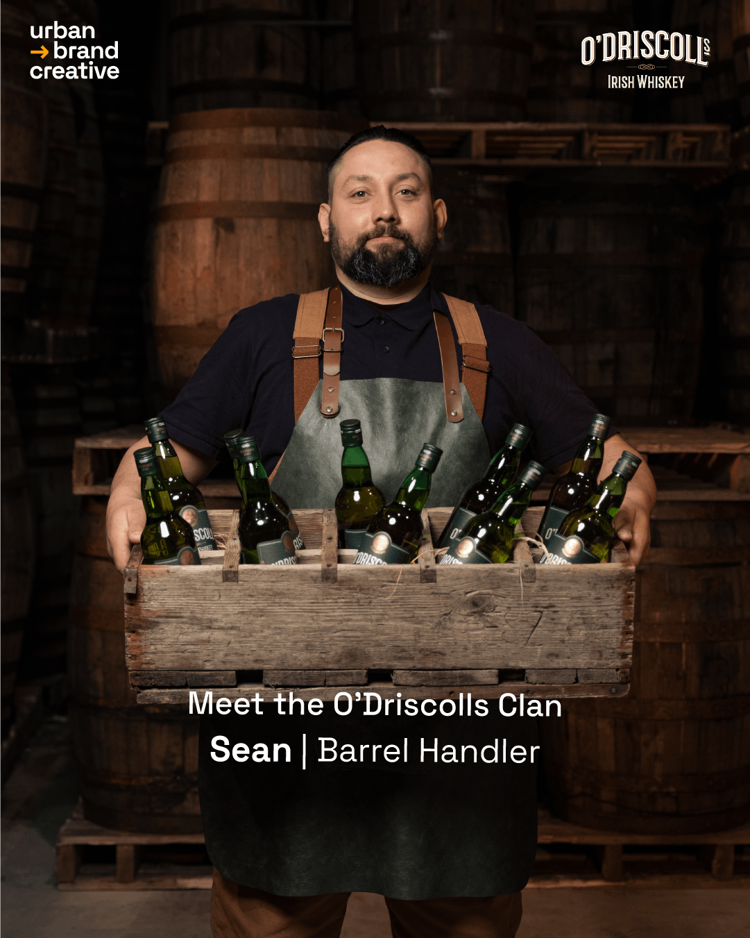 A man with a beard holding a wooden crate filled with green beer bottles in a cellar with wooden barrels in the background.