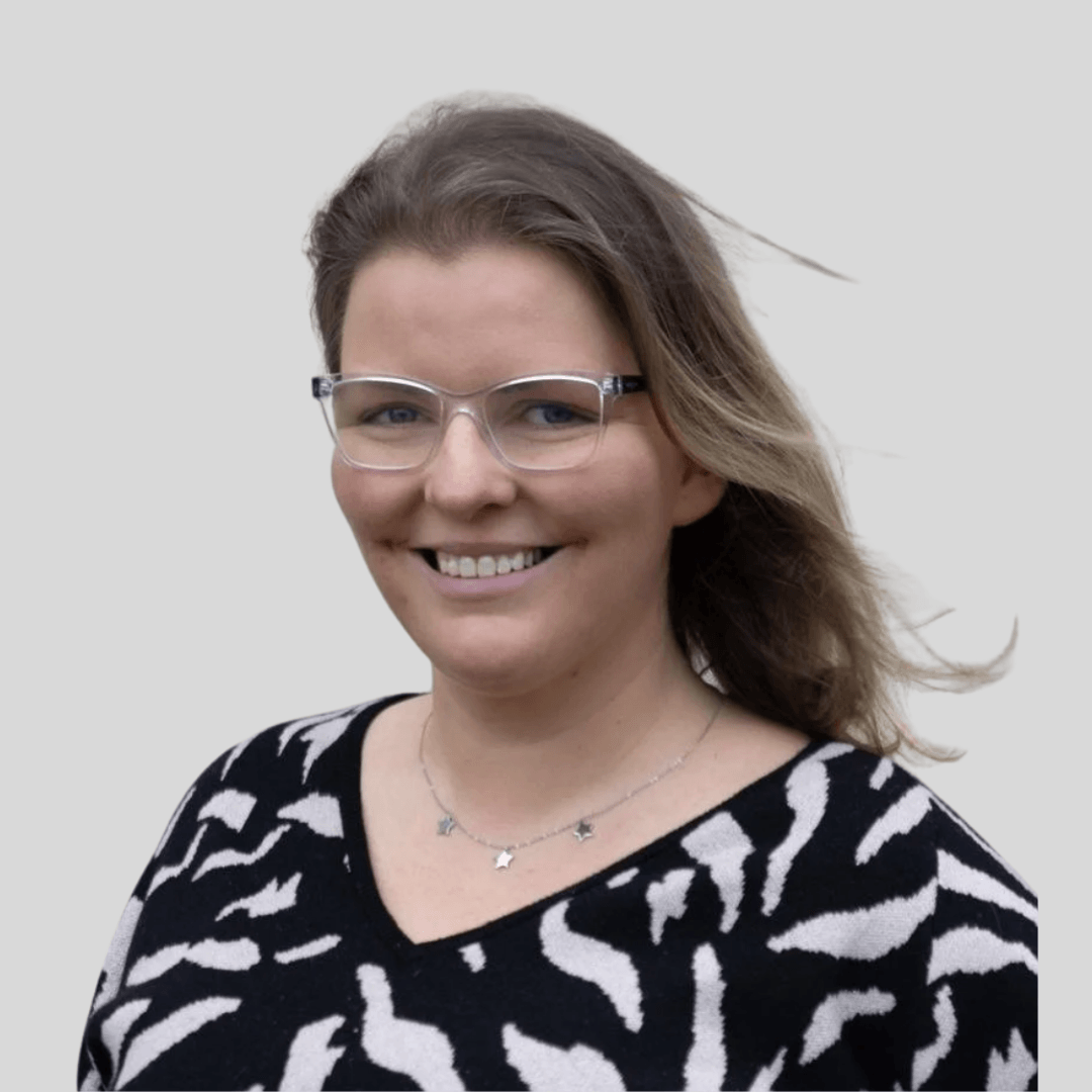 A woman with shoulder-length brown hair, wearing clear glasses, a black and white animal print top, and a silver necklace, smiling against a plain light background.