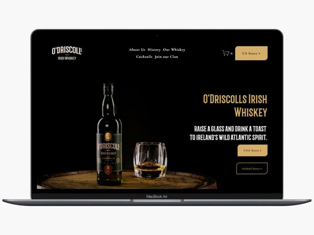 A website displayed on a laptop screen promoting O'Driscolls Irish Whiskey, featuring a bottle of whiskey, a glass with whiskey, and navigation options for different stores.