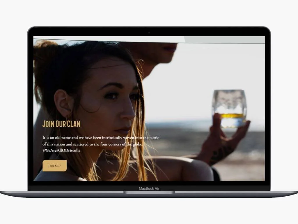 A woman with long hair and tattoos holding a glass of whiskey at the beach, with a man partially visible behind her, on a MacBook Air screen.