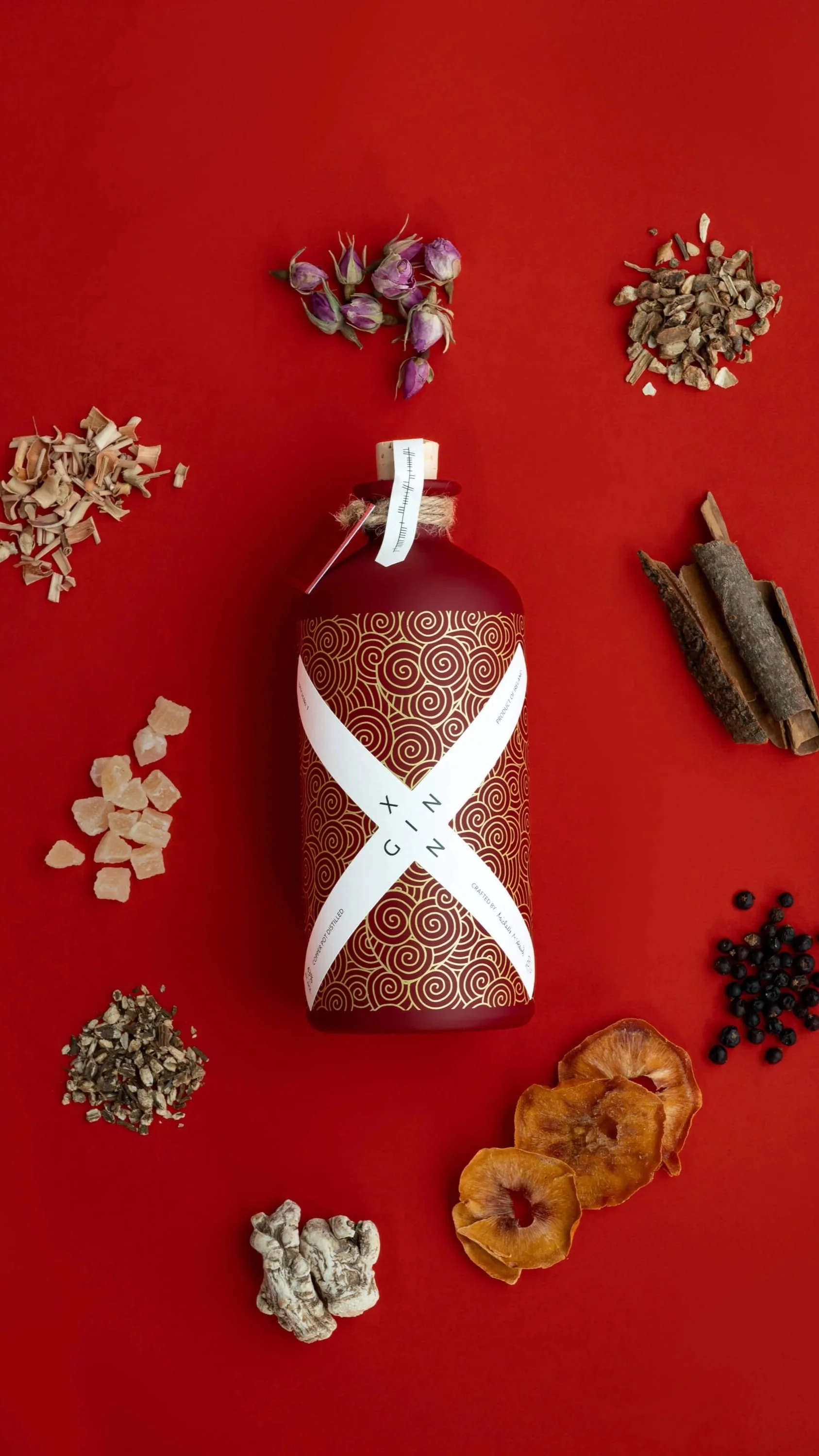 A red bottled tonic surrounded by various herbs and spices on a red background.