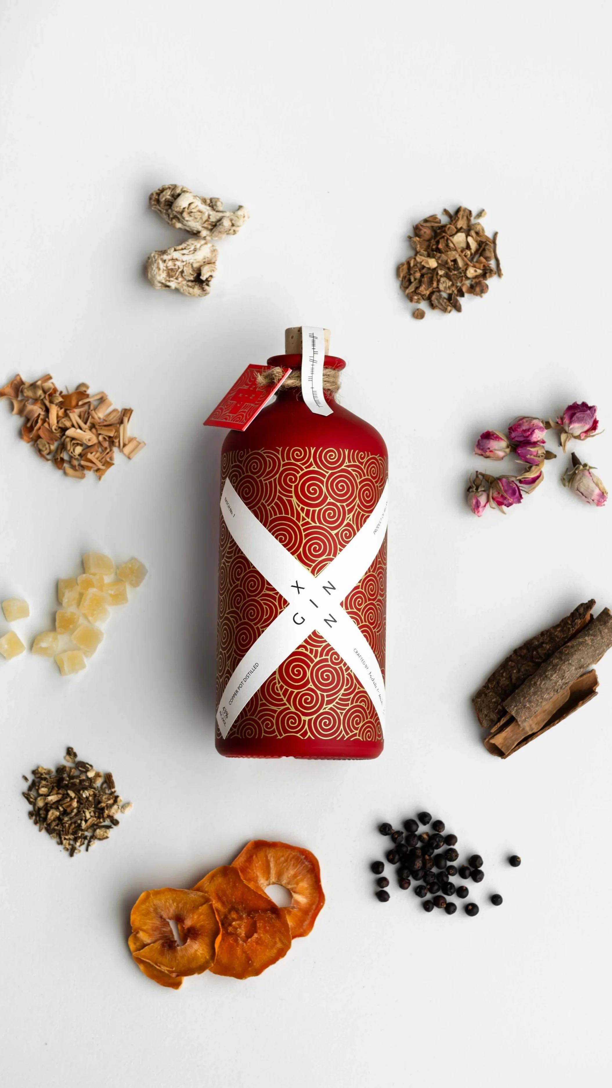 A red bottle with gold swirl patterns and a white ribbon label surrounded by various dried herbs, flowers, and spices on a white background.