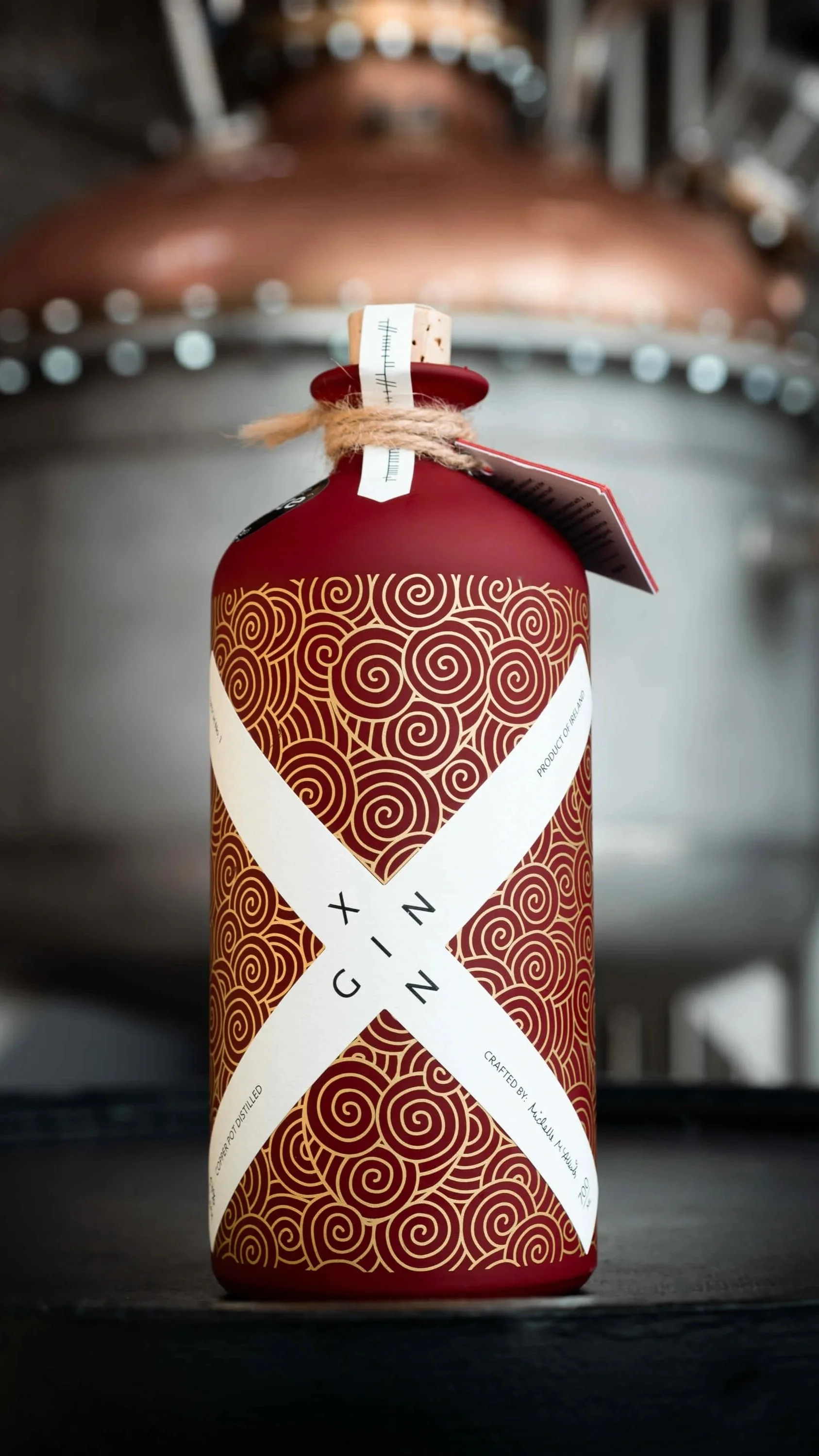 A bottle of ginger-infused alcohol with a white crisscross label, placed on a dark surface with metallic fermentation tanks in the background.