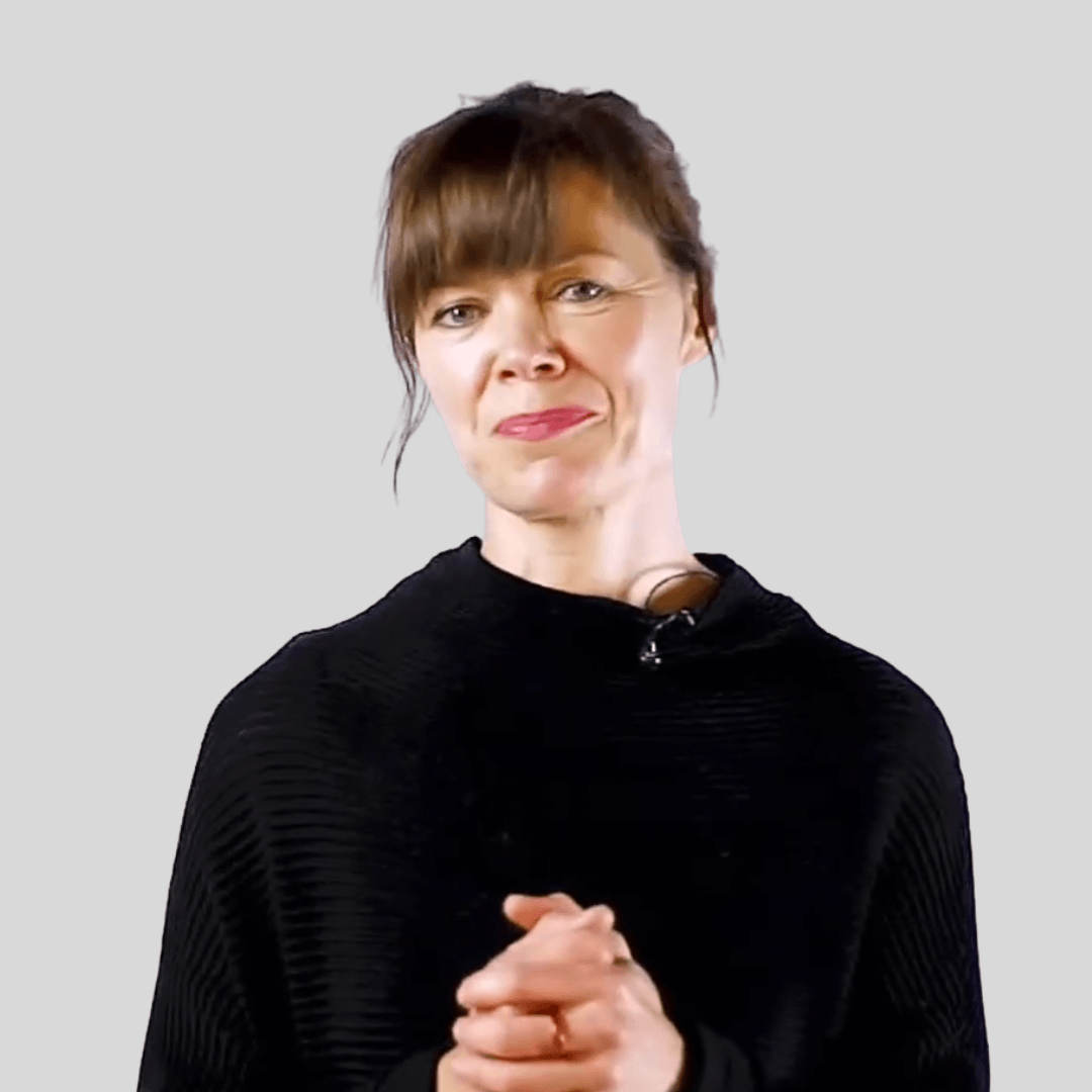 A woman with short brown hair and wearing a black top, standing against a plain light gray background, looking directly at the camera with a slight smile, clasping her hands together.