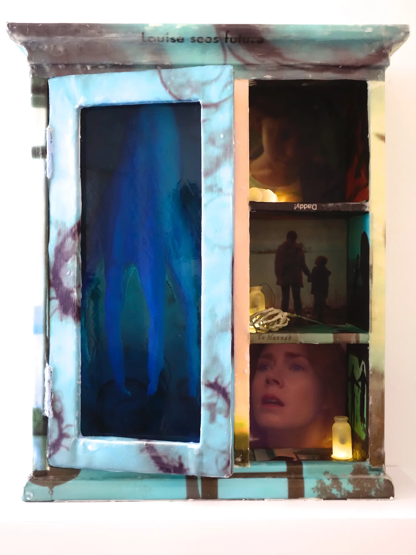 Arrival, 2018, 20”x24”x9”, screen grabs from the film printed on paper and attached to a wall cabinet with encaustic. Branch, bottles, lights and a toy animal. $2,000