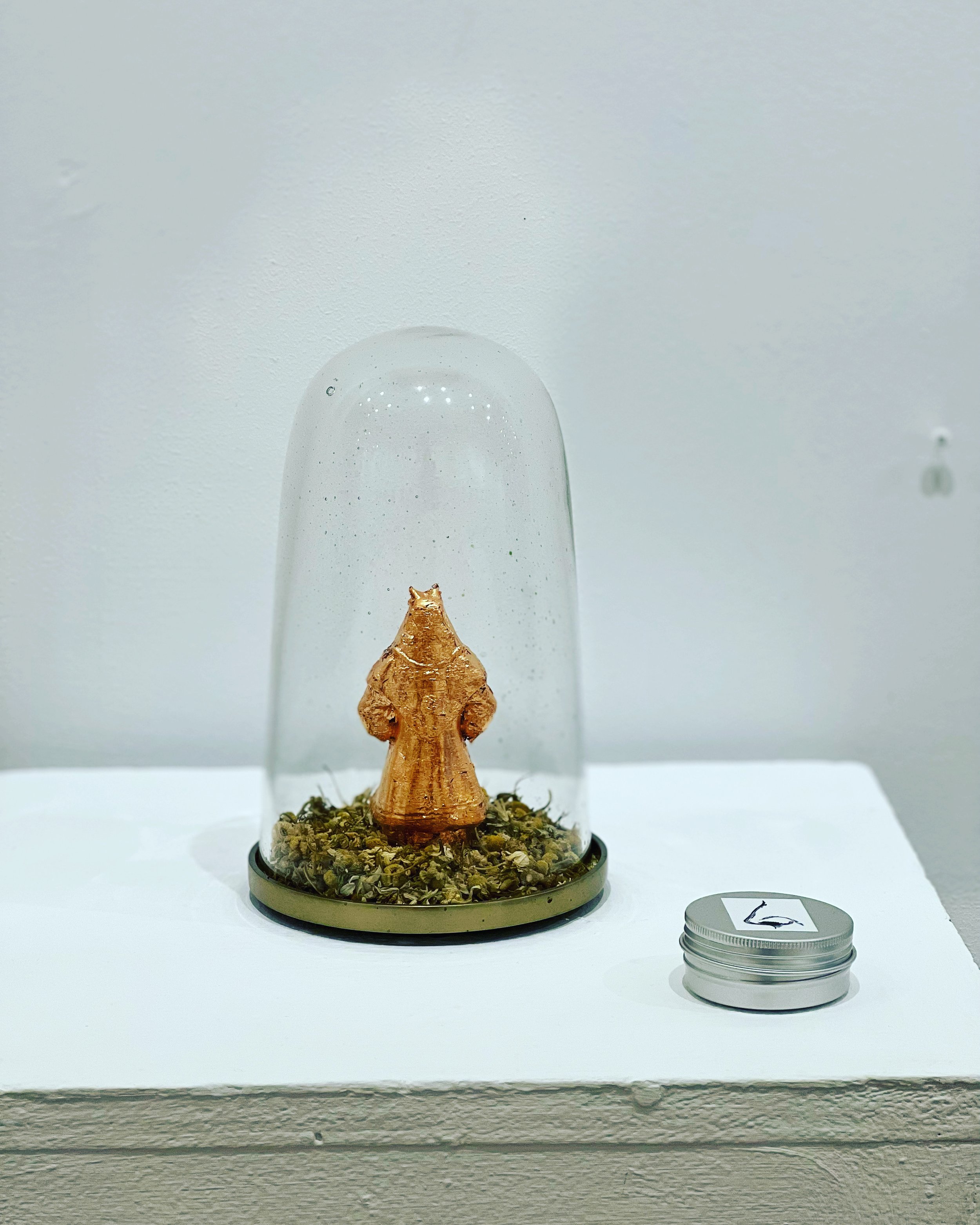   Back Story , 2022  3D printed figurine, copper gilding, chamomile, chamomile oil, brass stand, glass cloche  5 x 8 inches   $750.   A 3-D printed double-sided figurine, gilded with copper, sitting on a layer of chamomile flowers and displayed in a 