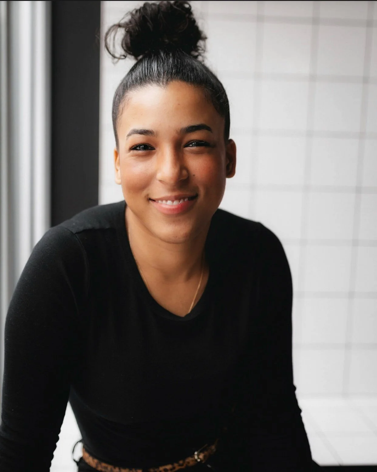 A woman with dark curly hair tied up in a bun, wearing a black top, smiling while looking at the camera indoors with white tiled walls in the background.